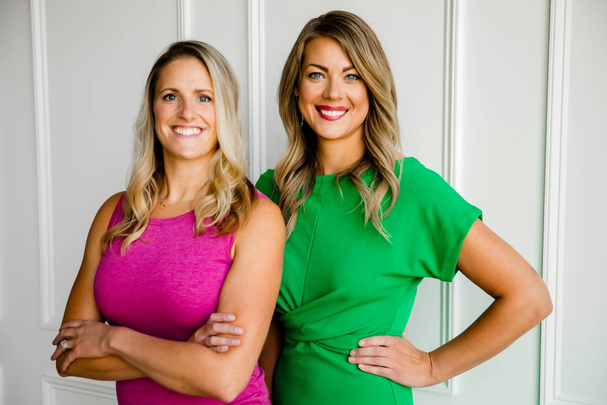 Two women smiling and posing together indoors, one wearing a sleeveless pink top with arms crossed, the other in a bright green dress with one hand on her hip.