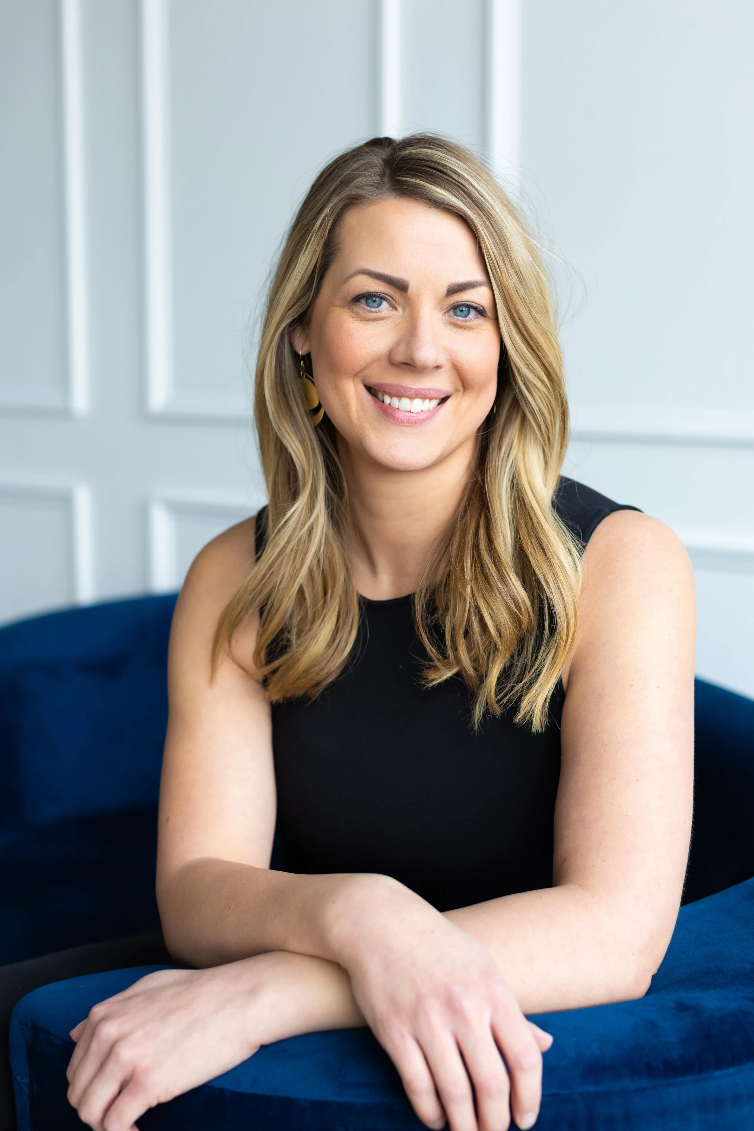 A woman with blonde hair, blue eyes, wearing a black sleeveless top, smiling, sitting with her arms crossed on a blue sofa in a bright room with white paneled walls.