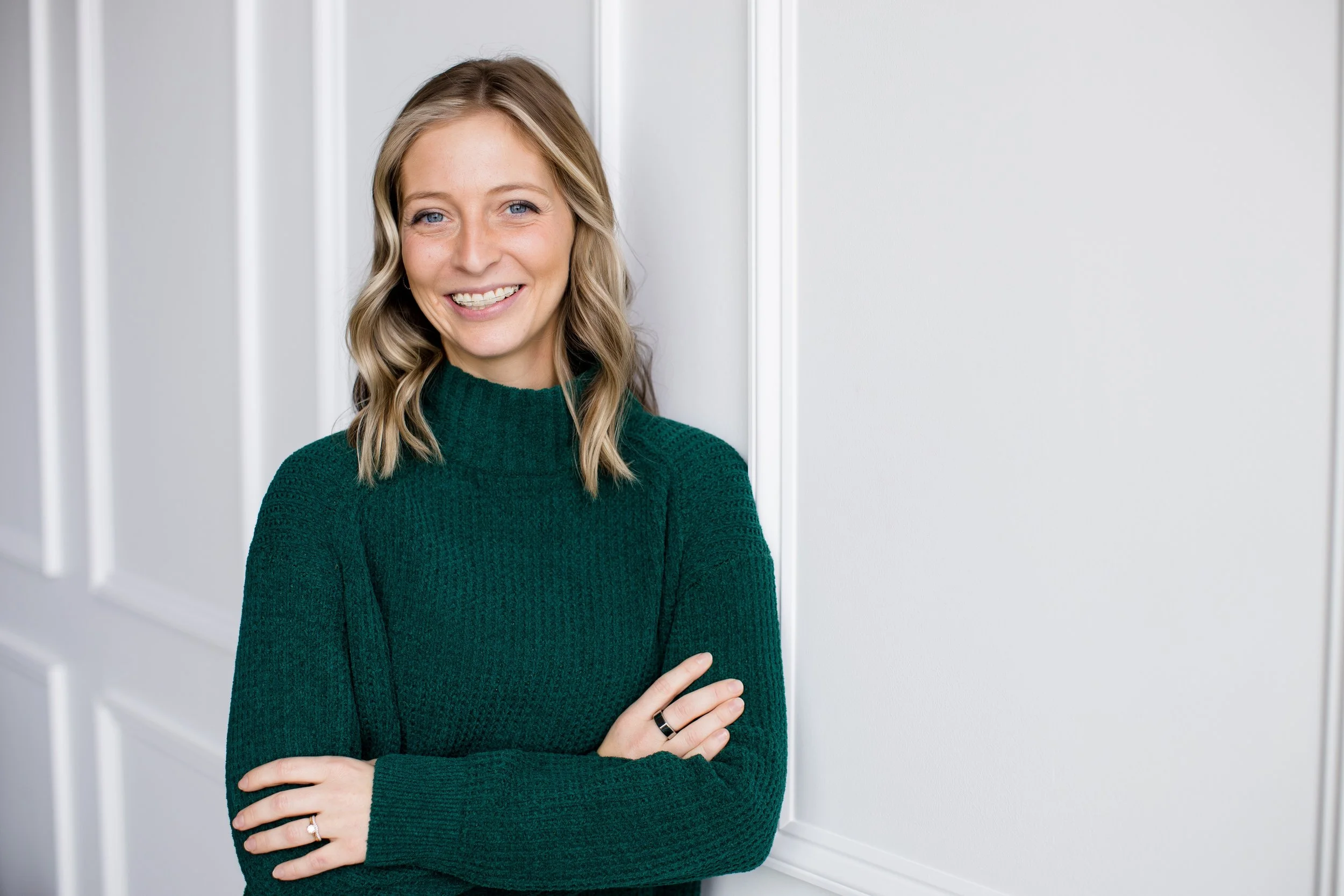 A woman with blonde hair wearing a green sweater, standing with her arms crossed and smiling, indoors with white walls.