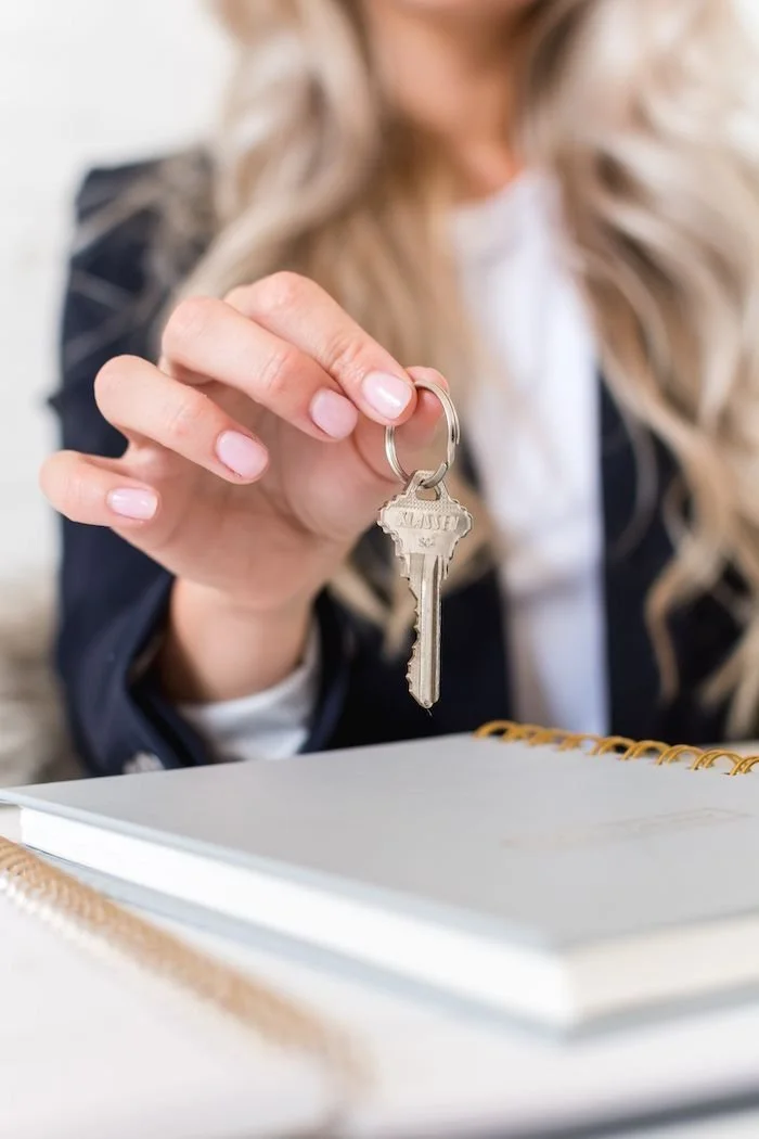 A woman with long, blonde hair holding a silver key attached to a keyring, sitting at a desk with a notebook or planner in front of her.