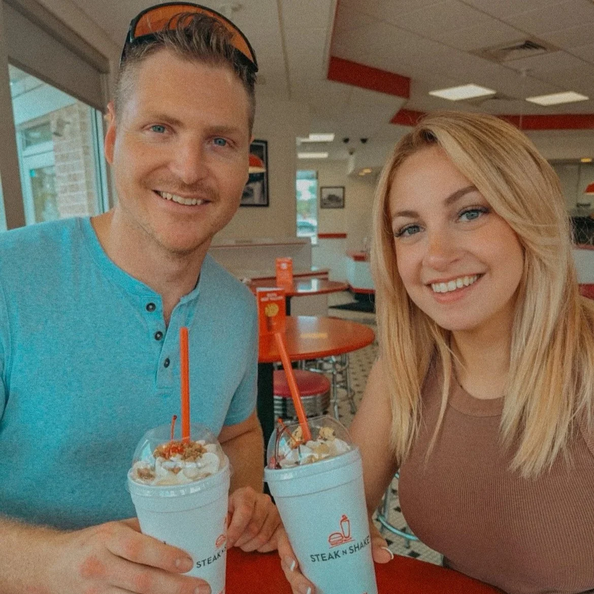 A man and woman smiling at a diner, each holding a milkshake with whipped cream and caramel toppings.