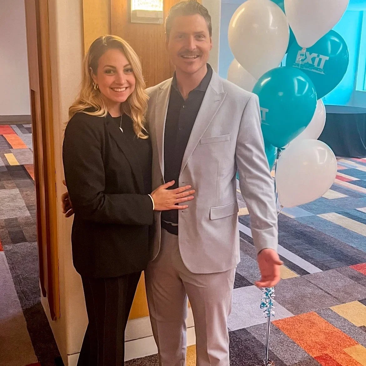A woman and a man smiling and posing together at an event, with balloons in the background.