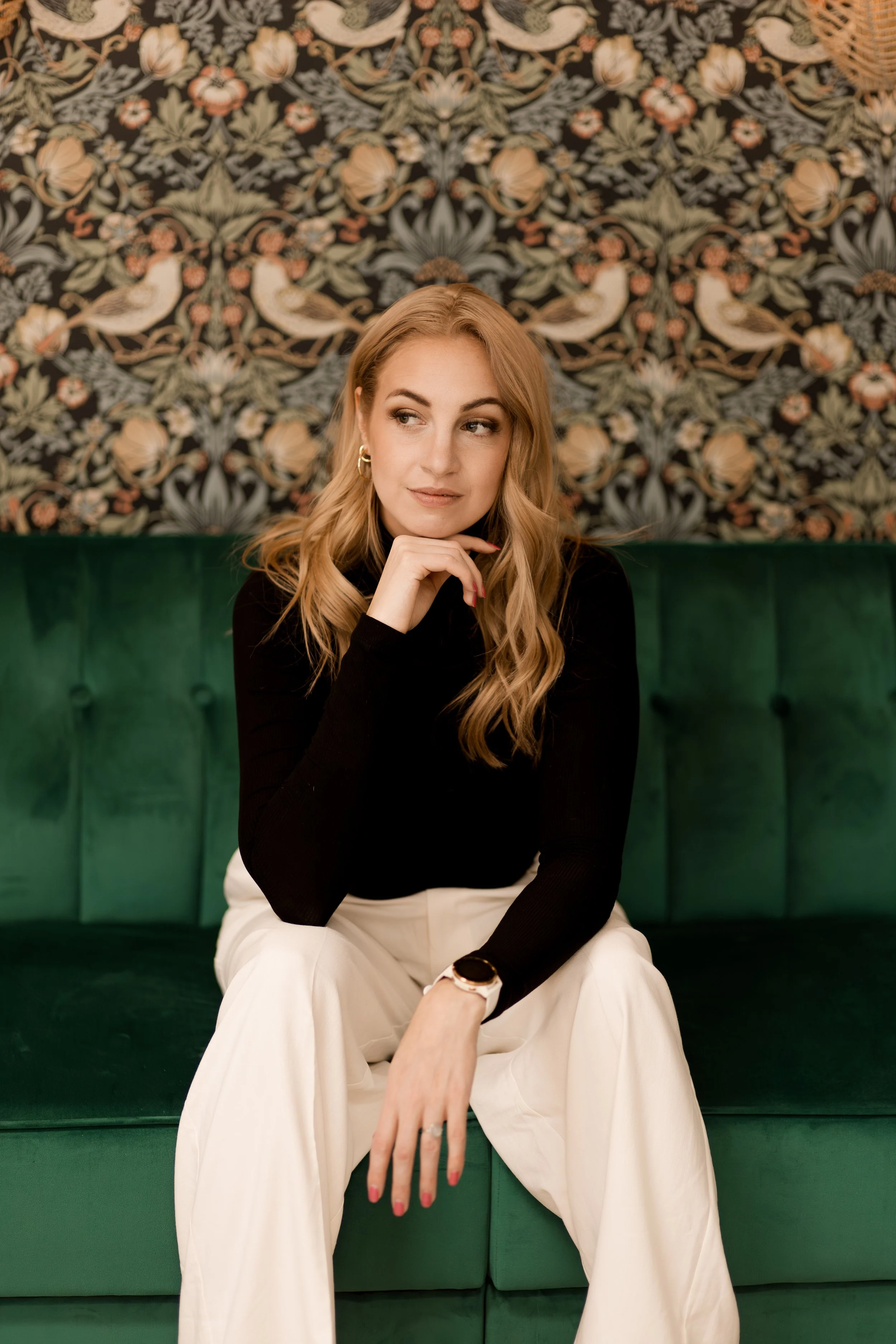 A young woman with blonde hair sitting on a green velvet sofa in front of a decorative floral wallpaper with birds.