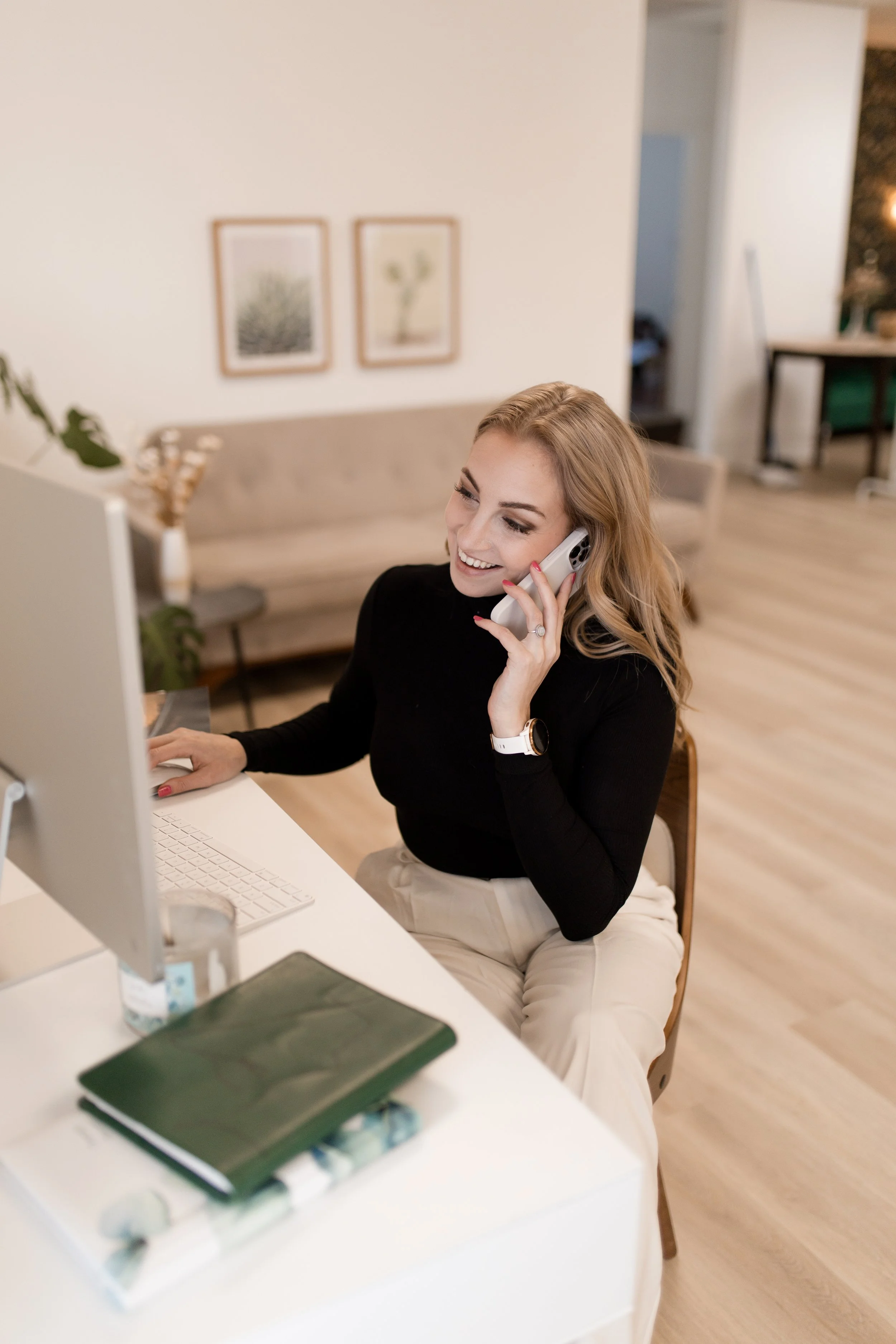 A woman with blonde hair, wearing a black top and cream pants, sitting at a desk, talking on the phone and using a computer.