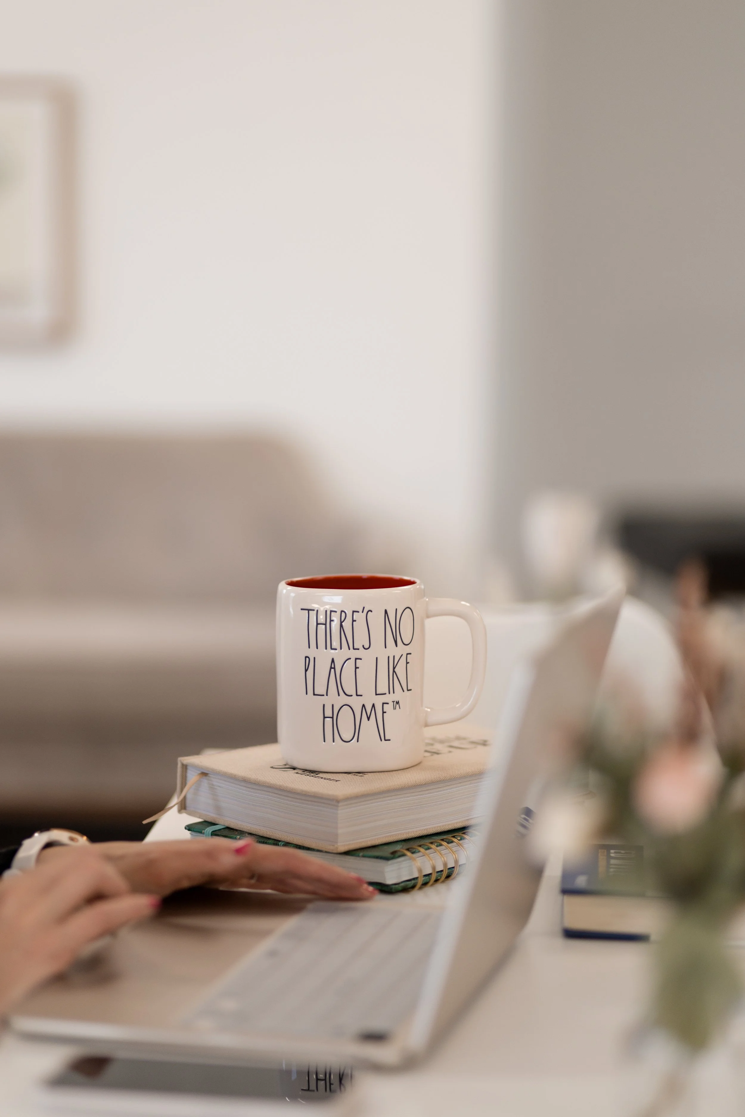 A white coffee mug with the text 'There’s no place like home' on it, resting on top of a closed book, on a desk next to a person using a laptop in a cozy indoor setting.