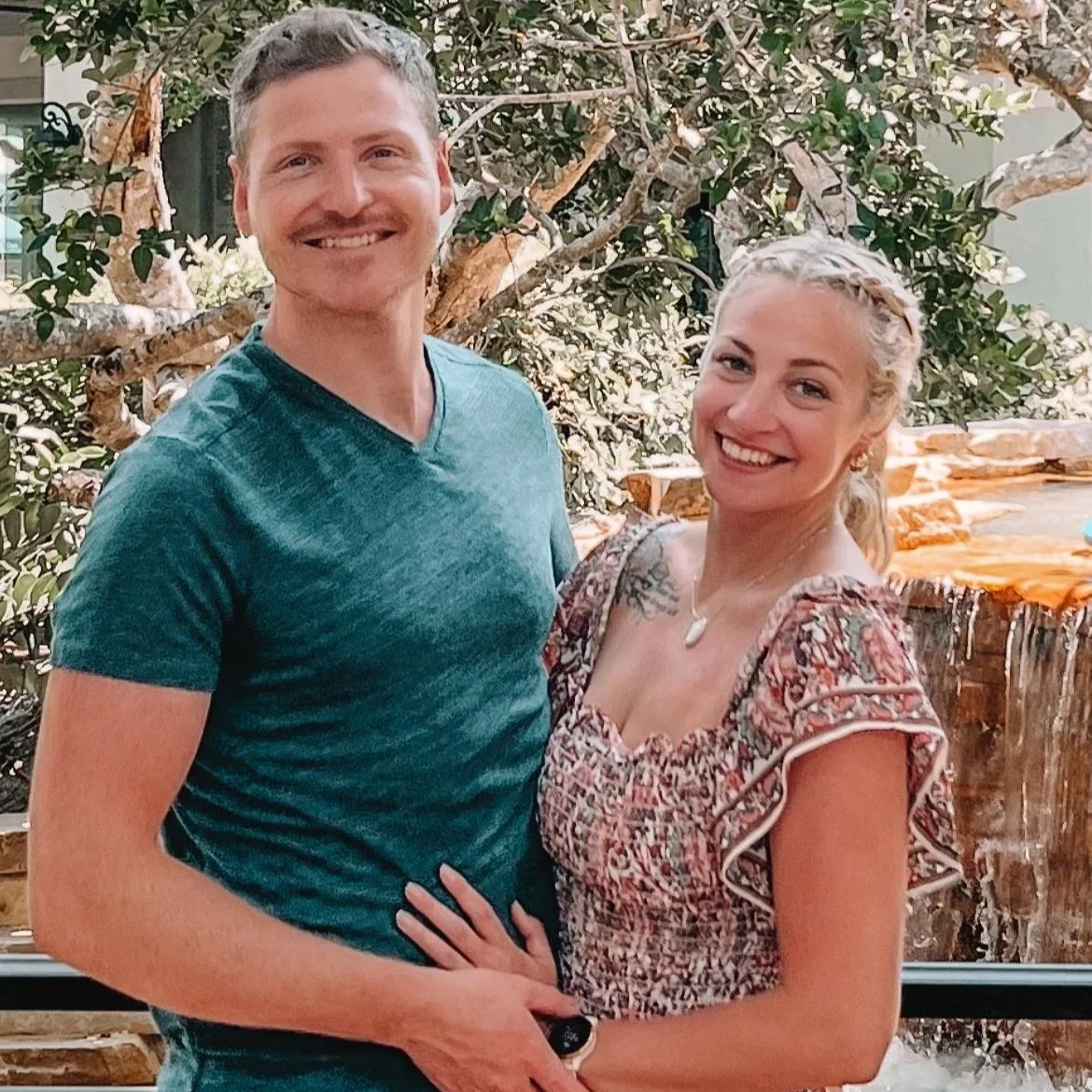 A smiling man and woman standing close together outdoors in front of a tree and a wooden water feature, embracing each other.