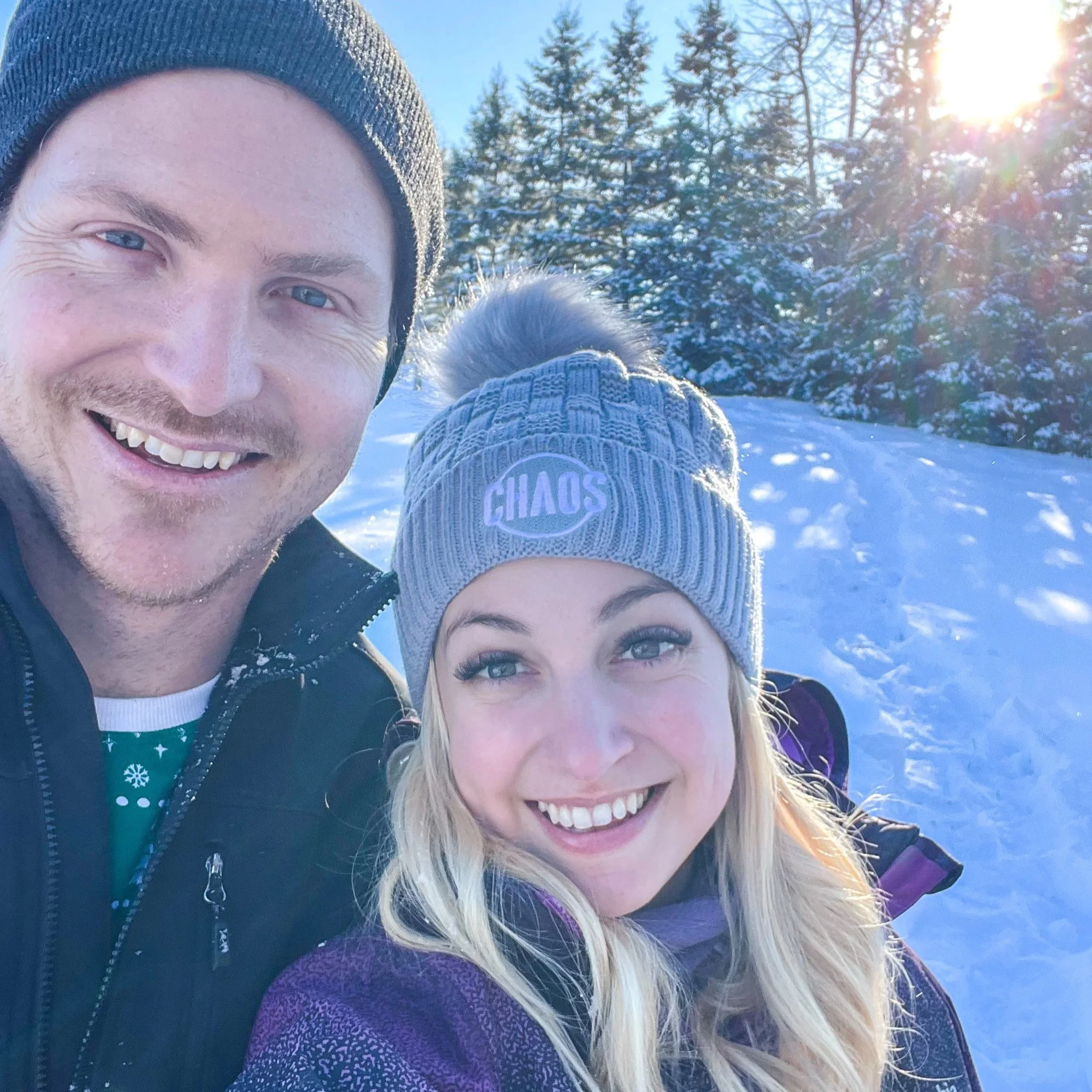 A man and woman smiling outdoors in a snowy landscape with trees and sunlight in the background.