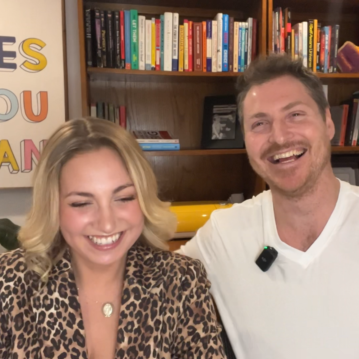 Two smiling people, a woman with blonde hair wearing a leopard print top and a man with brown hair and a beard wearing a white shirt, sitting together in front of a bookshelf in a room.