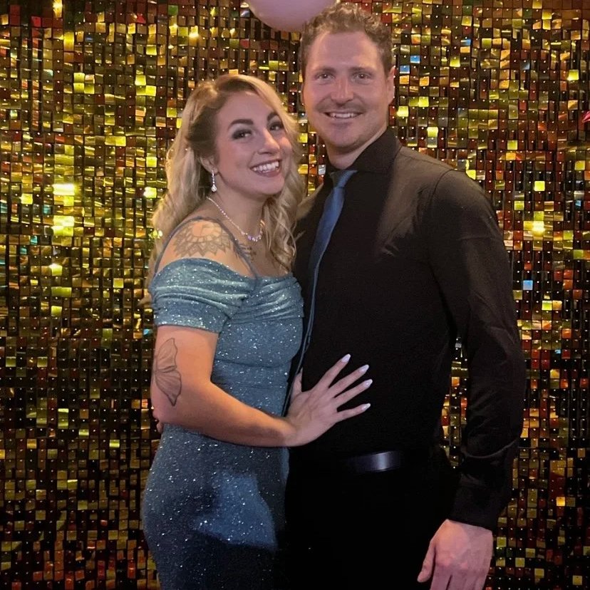 A smiling couple, a woman in a sparkling blue off-the-shoulder dress and a man in a black shirt and tie, standing close together at a party or celebration with a shiny, golden, mosaic-like background.