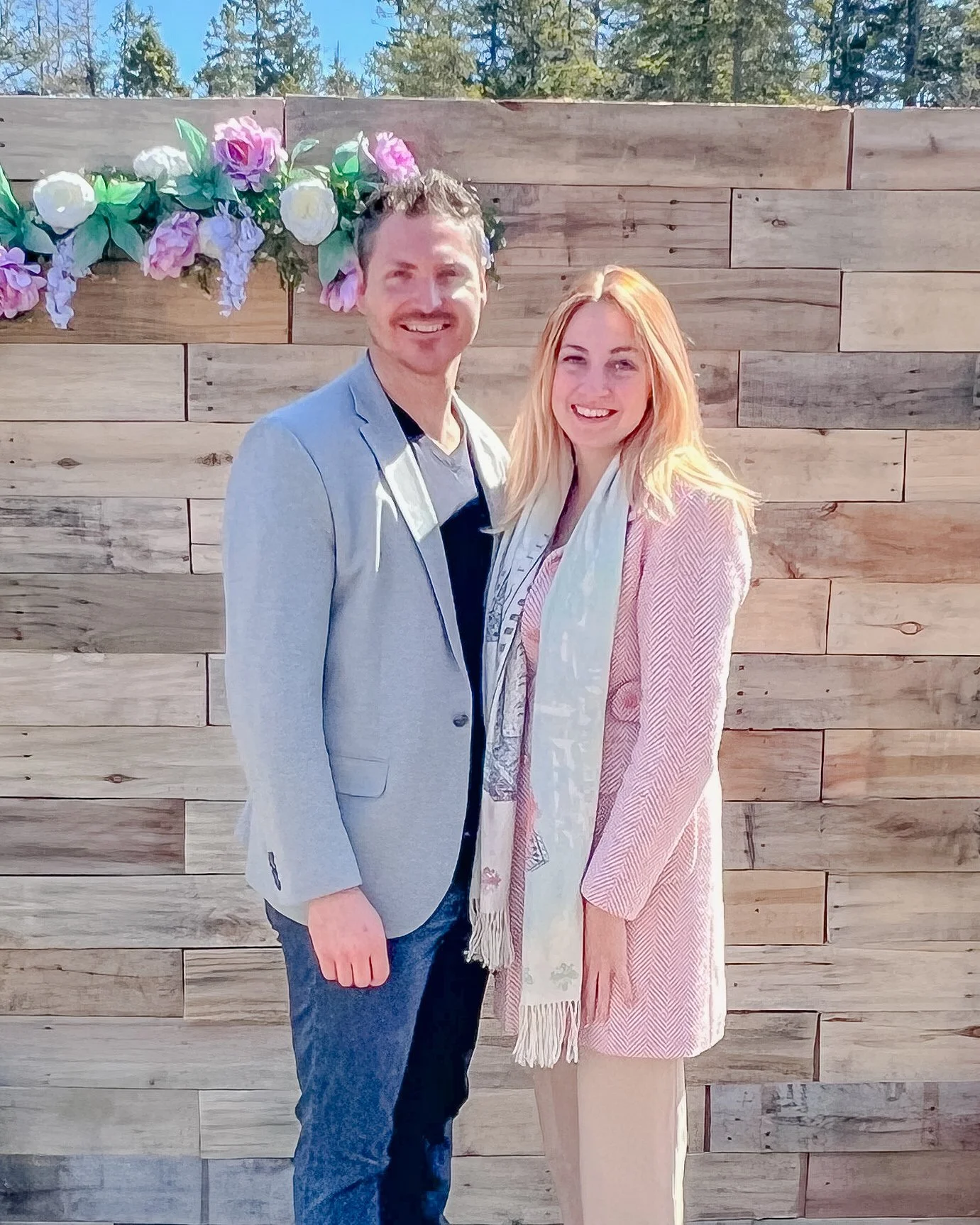 A man and a woman smiling and standing close together outdoors in front of a wooden wall decorated with pink and purple flowers.