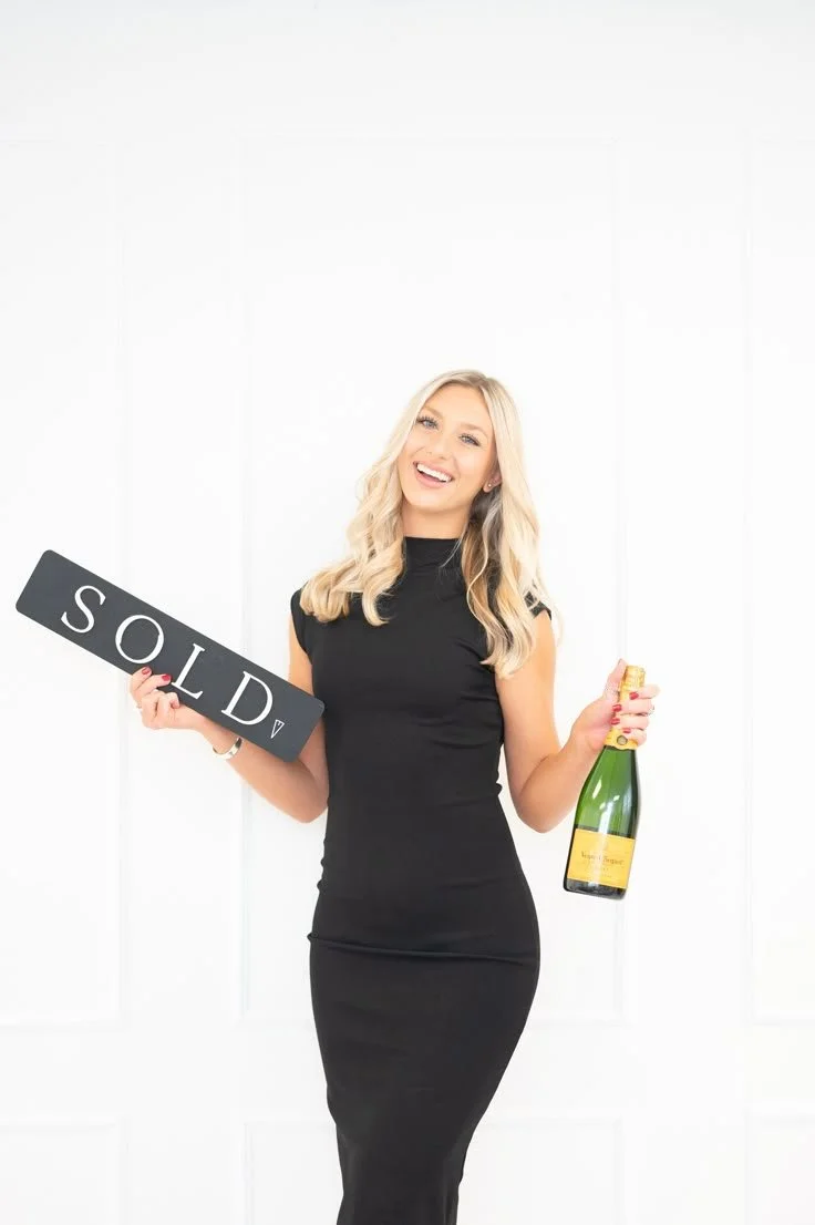 A woman in a black dress holding a 'SOLD' sign and a bottle of champagne, smiling happily.