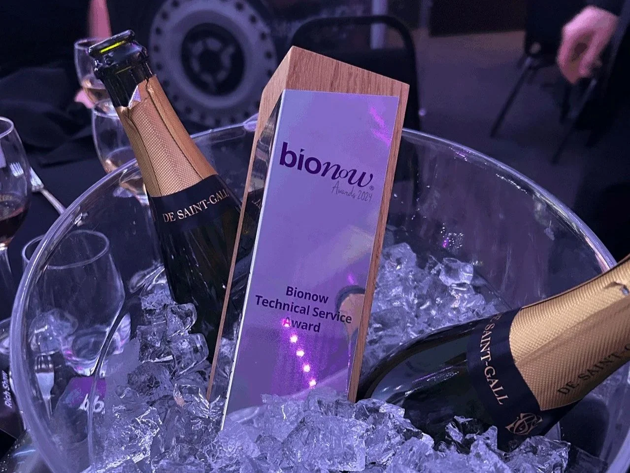 A clear ice bucket holding two bottles of champagne and a framed award for Bionow Technical Service Award 2024