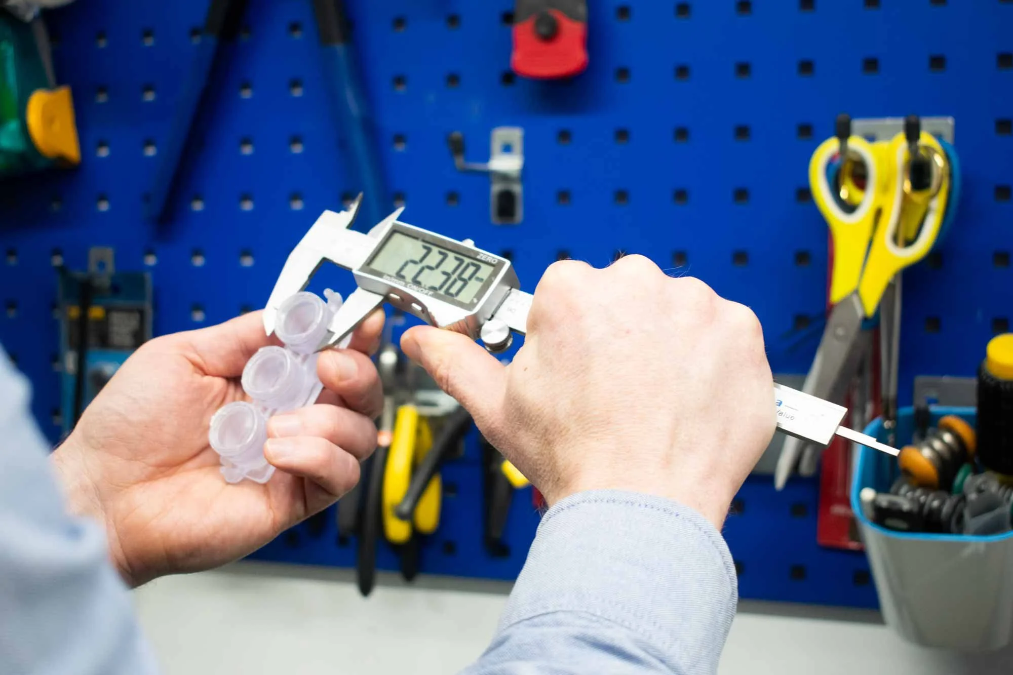 Person using a digital caliper to measure a small object in a workshop, with tools and scissors on a blue pegboard in the background.