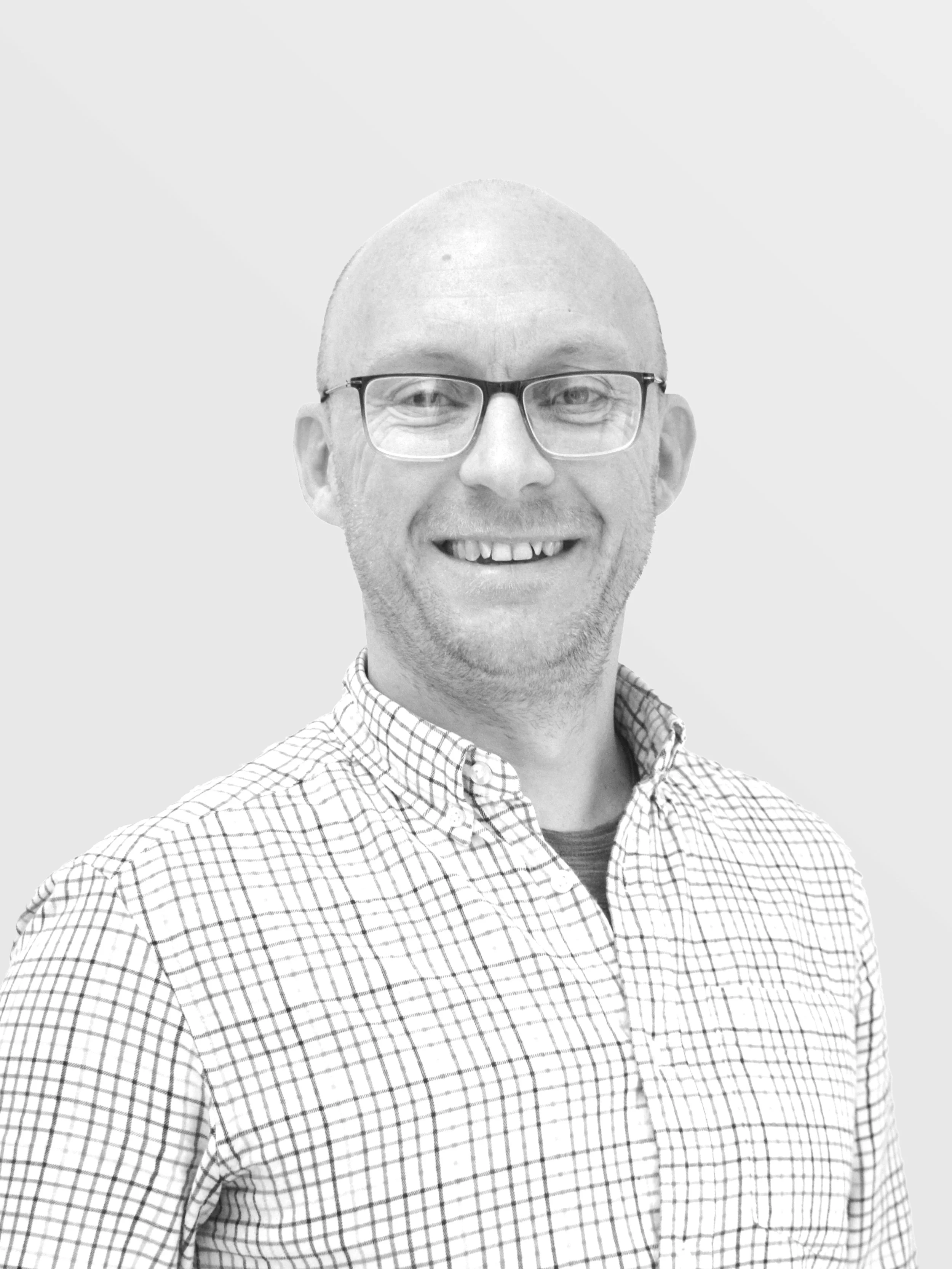 Black and white portrait of a smiling man with glasses, a checkered shirt, and short hair, standing against a plain background.