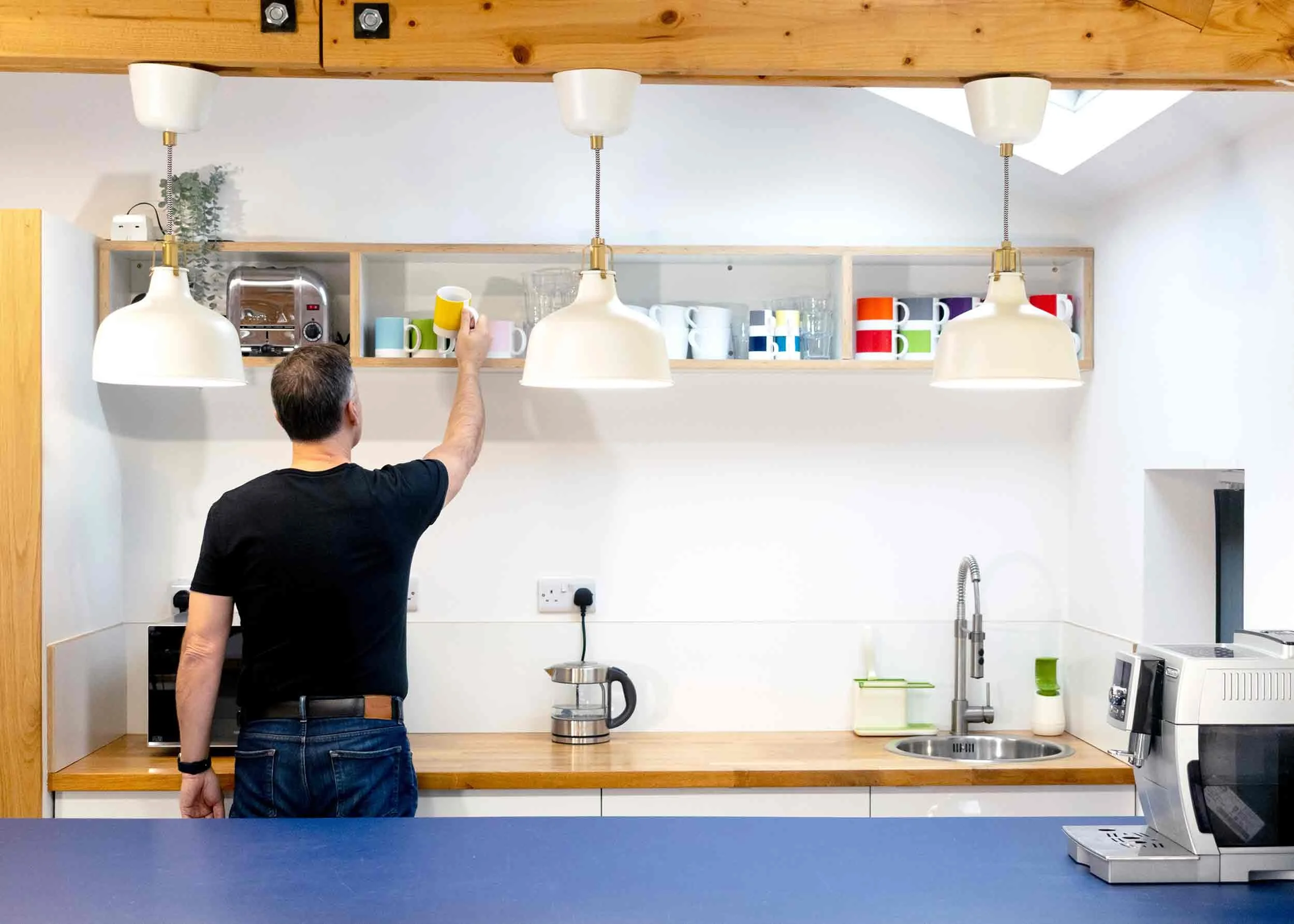 Man reaching to pick a colorful mug from a shelf in a modern kitchen with white and wooden surfaces, hanging lamps, and various kitchen appliances.