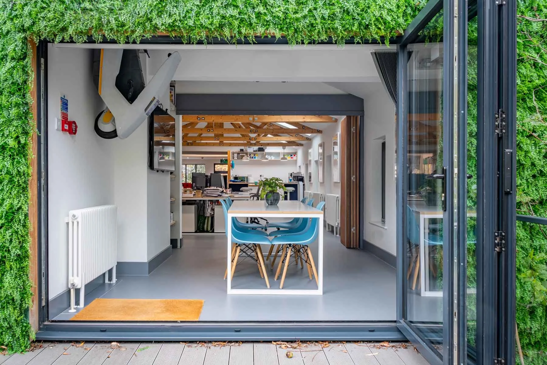 Open sliding glass doors reveal an office with a central table, blue chairs, a plant, and a monitor, with exposed wooden beams and various office equipment.
