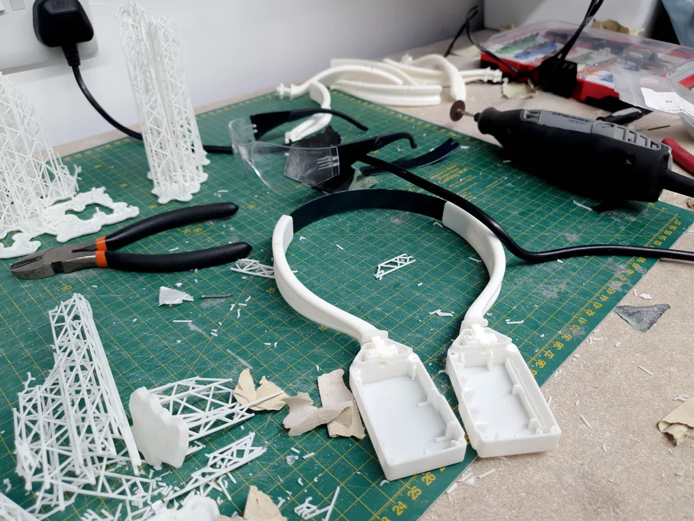 Disassembled 3D-printed structures and tools on a green cutting mat, including pliers, a drill, plastic parts, and safety glasses.