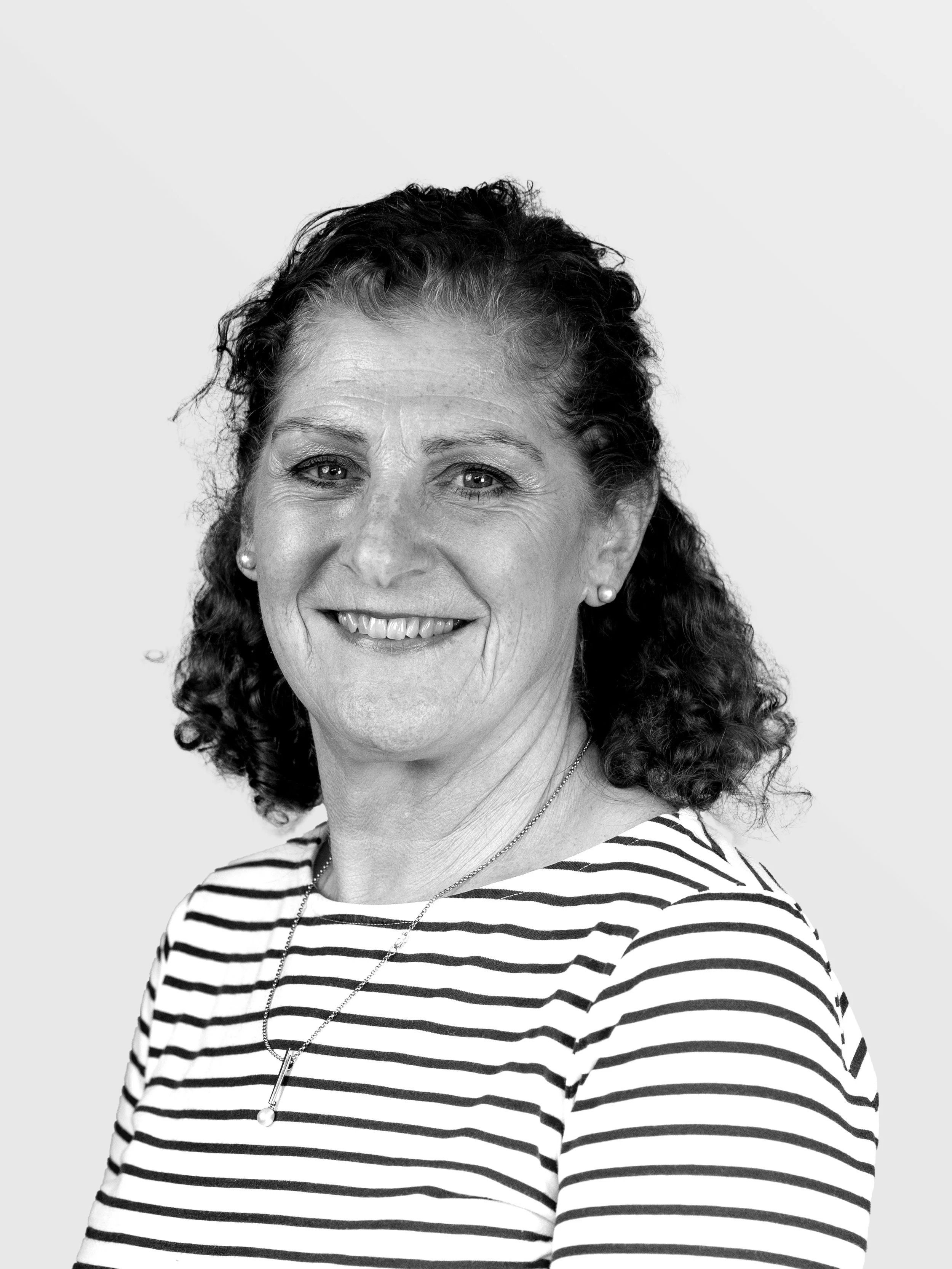 A smiling older woman with curly hair wearing a striped shirt and a necklace, against a plain background.