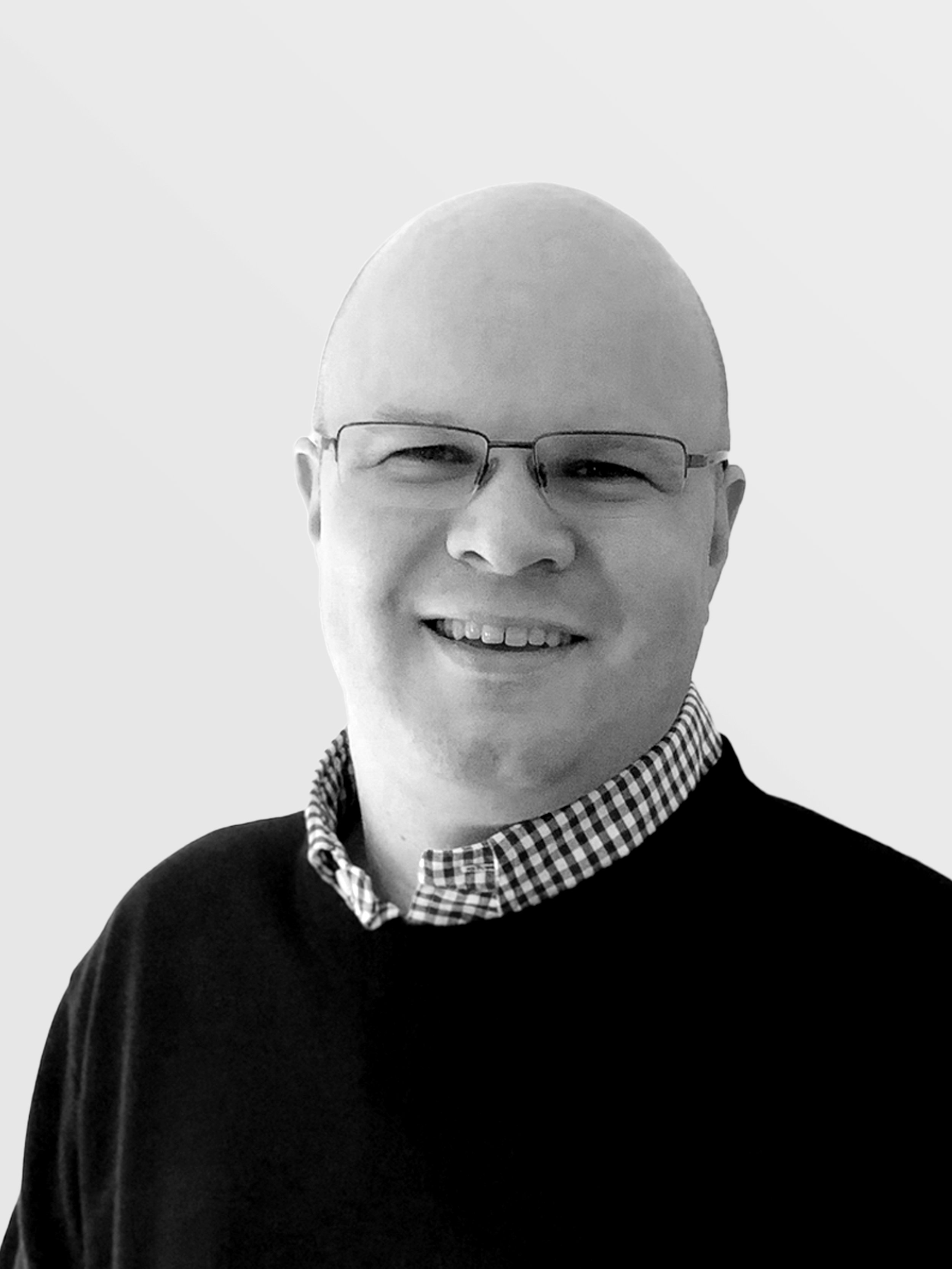 Black-and-white photo of a smiling man with glasses, wearing a checkered collared shirt under a dark sweater, against a plain white background.