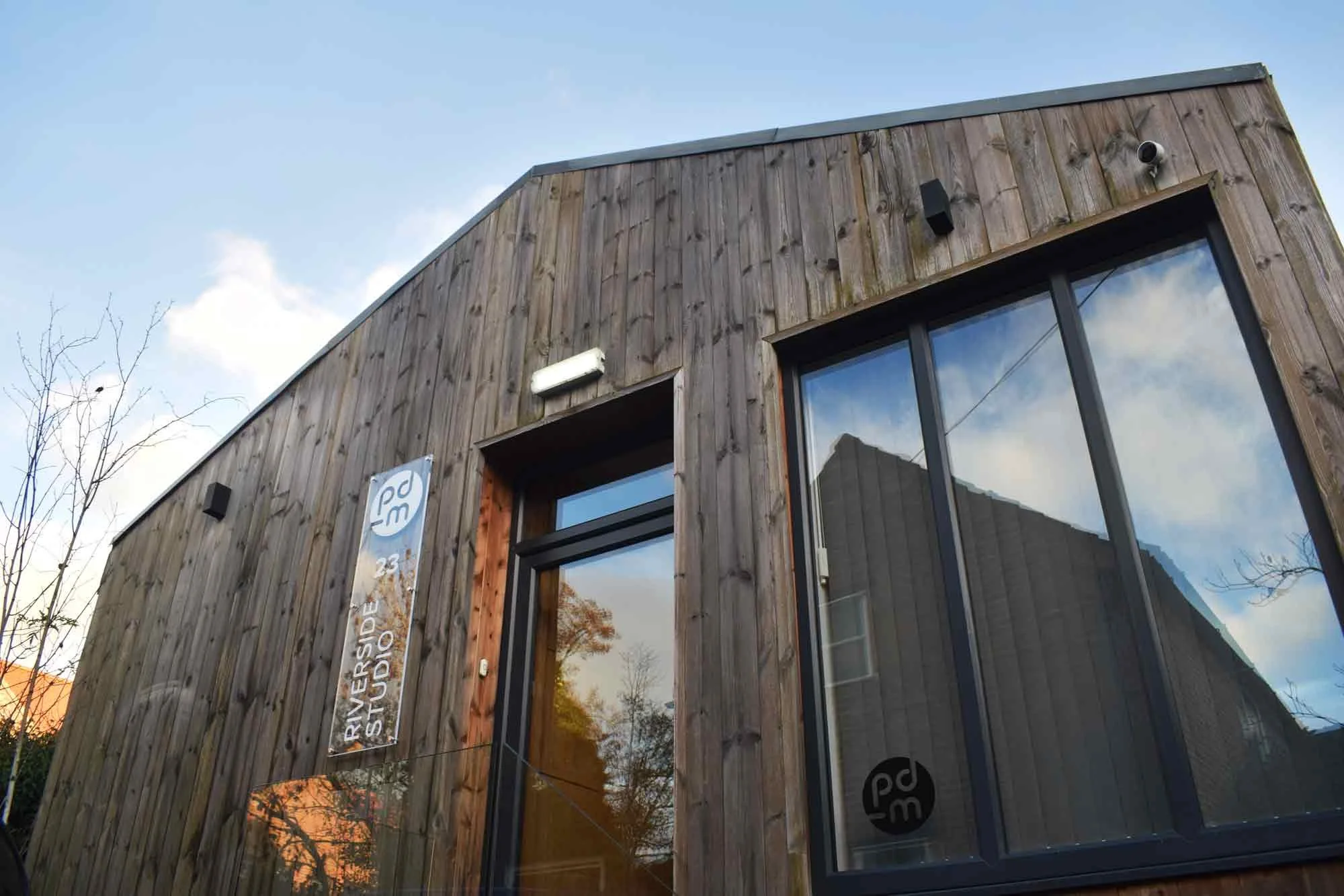Exterior of a modern building with a wooden facade, windows reflecting trees and the sky, with a sign that reads 'Riverside 23 Studio' and a logo next to it.