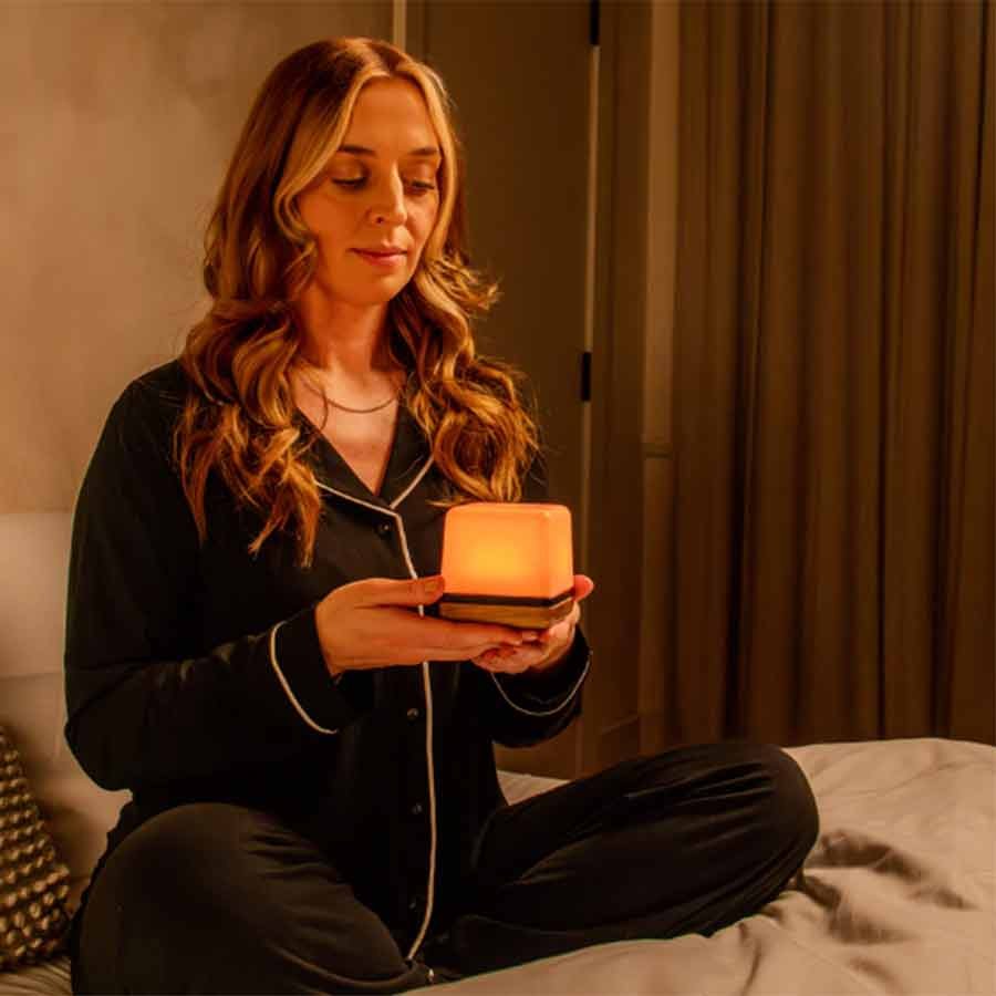 A woman sitting cross-legged on a bed, wearing black pajamas, holding a glowing salt lamp in her hands, in a cozy bedroom with warm lighting and beige curtains.