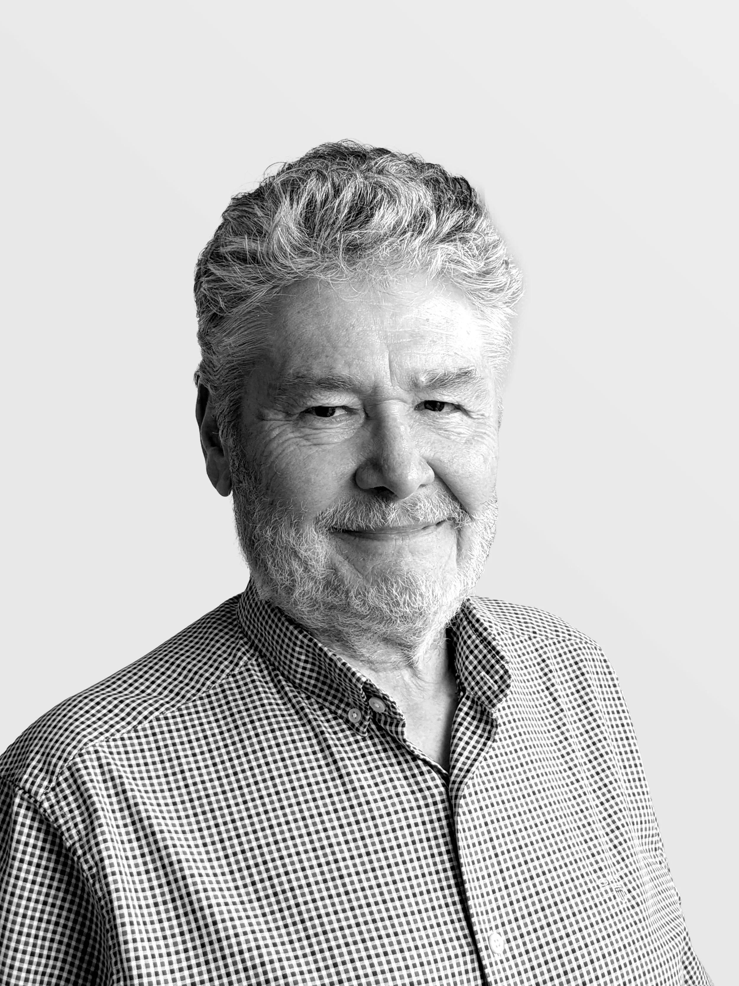Black and white portrait of an older man with gray hair and a beard, smiling slightly, wearing a checkered button-up shirt, against a light gray background.