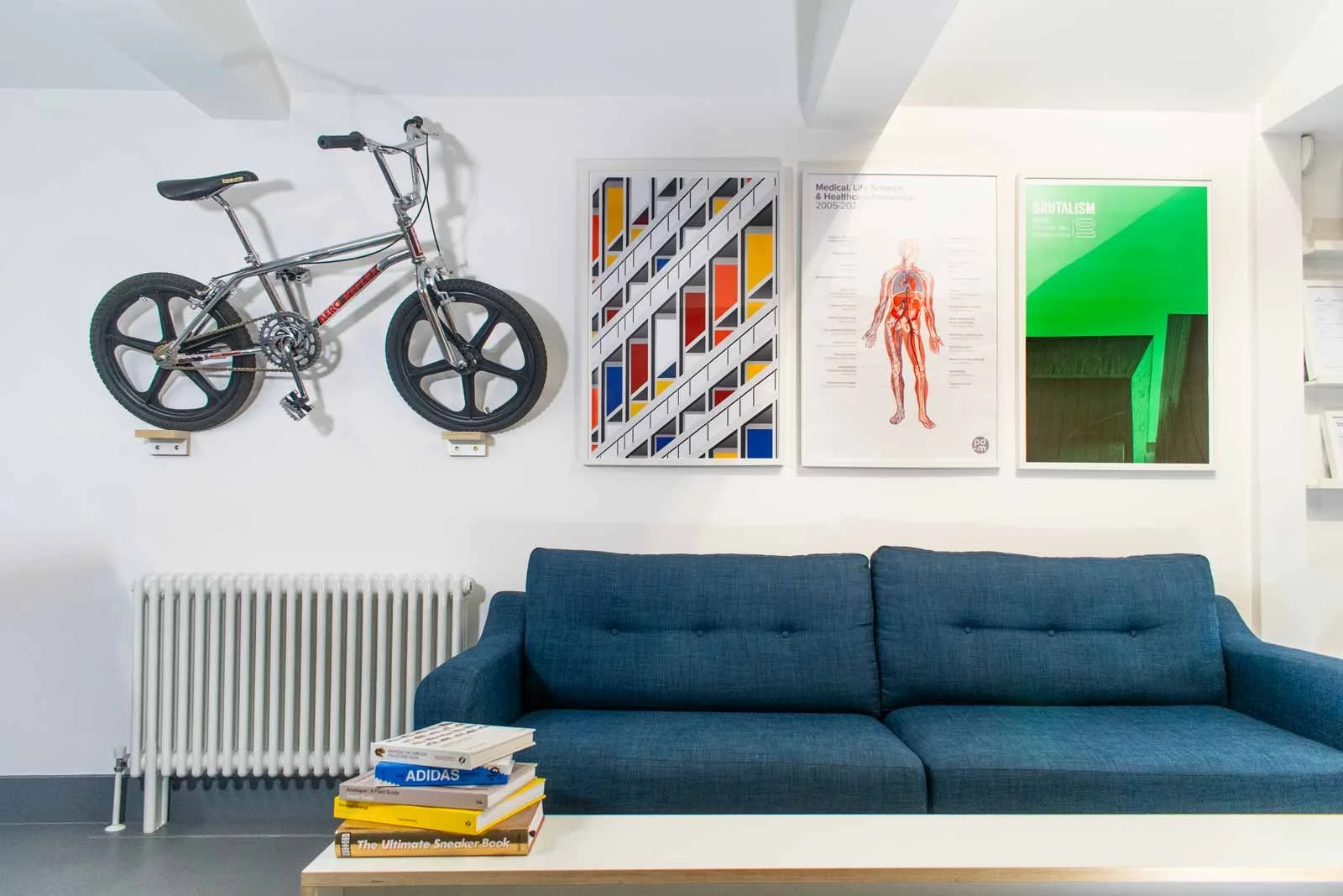 A white wall with a mounted bicycle, three colorful abstract art prints, a blue sofa, a white table with books including one titled 'The Ultimate Sneaker Book,' and a white radiator.