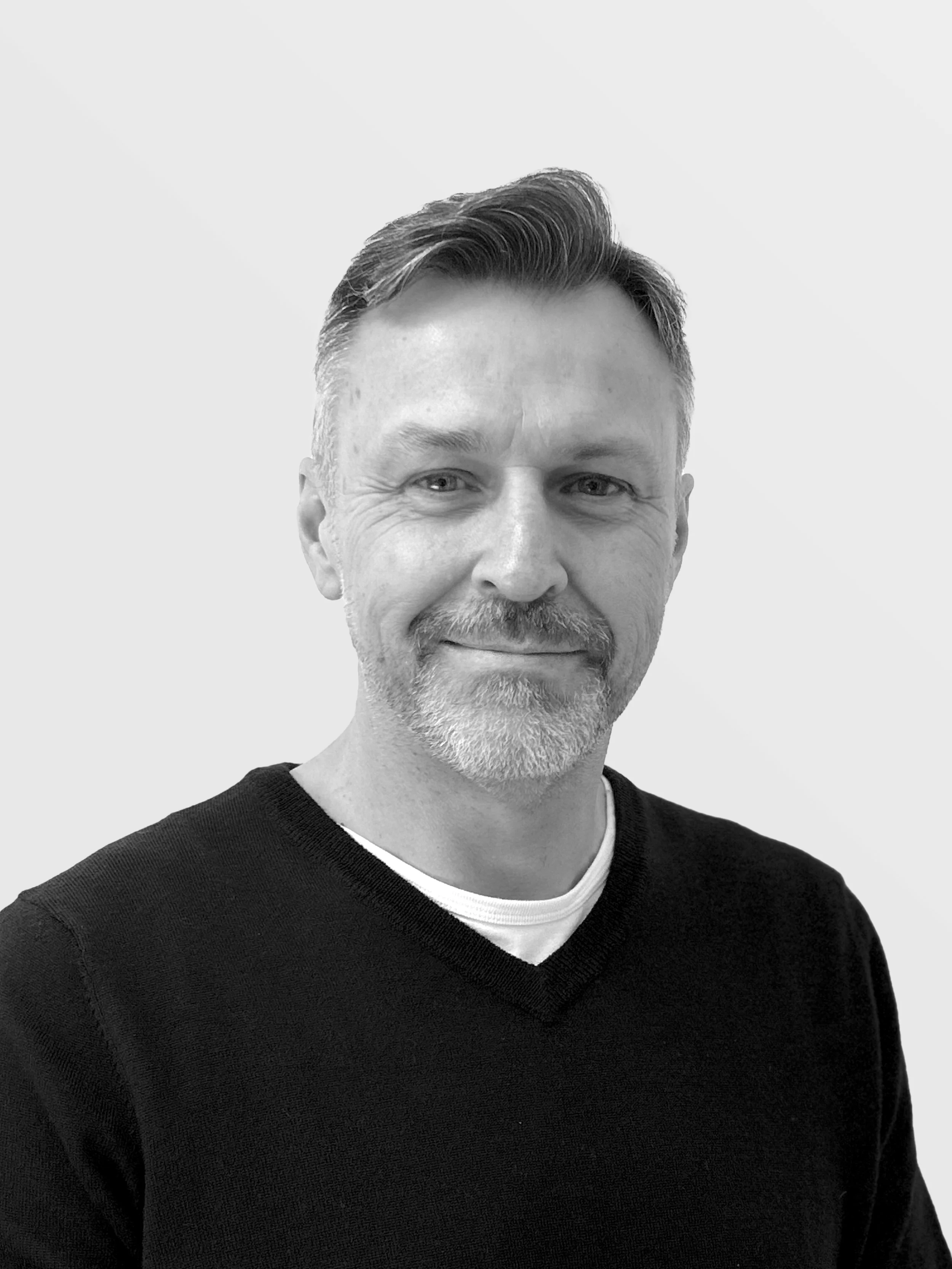 Black and white photo of a man with short, styled hair, a beard, and light eyes. He is wearing a black V-neck sweater over a white shirt and is smiling slightly. The background is plain and light-colored.