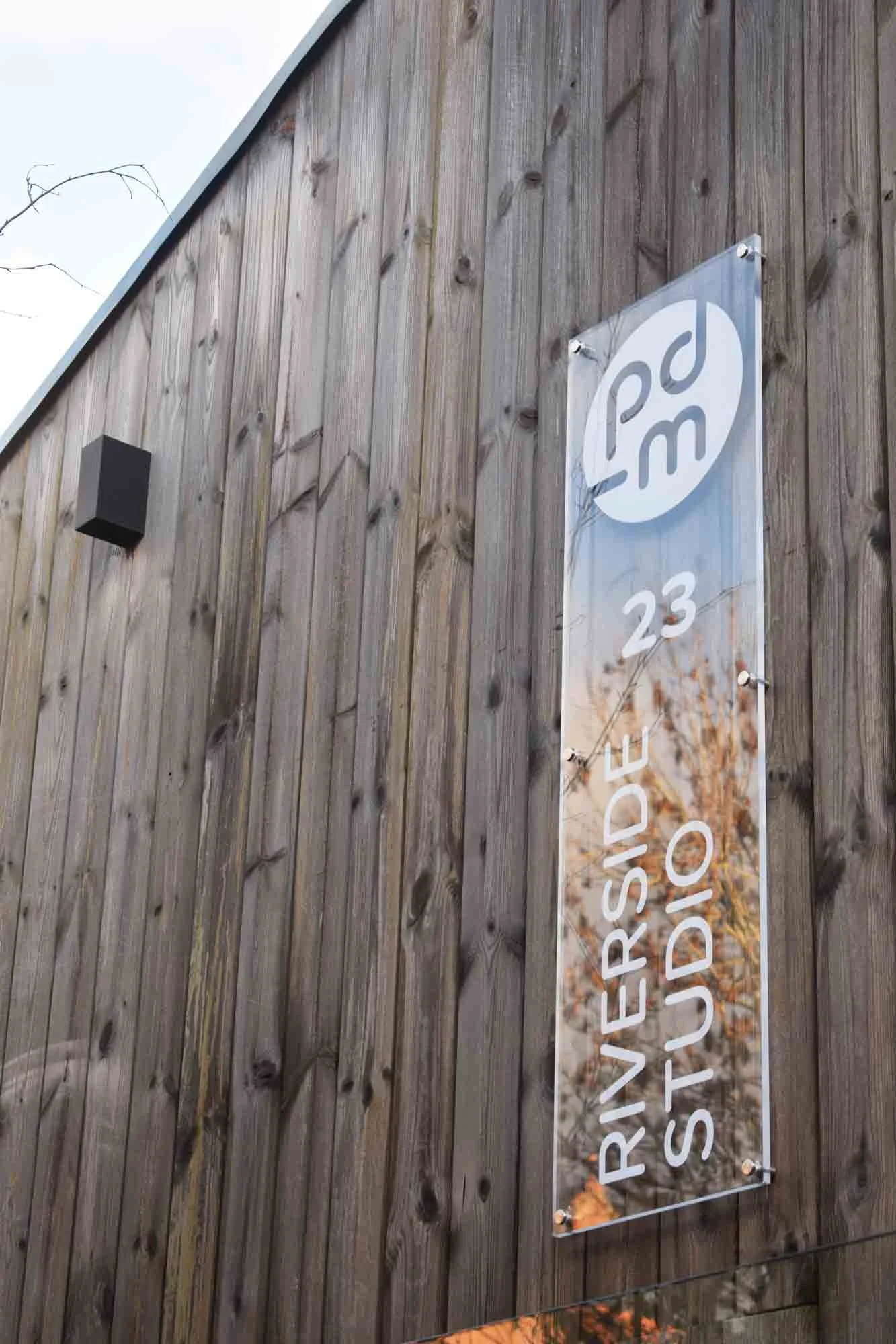 A vertical sign mounted on a wooden wall reads 'Riverside Studio 20,' with a circular logo above the text. The background shows a light blue sky, and there is a speaker mounted on the wall nearby.