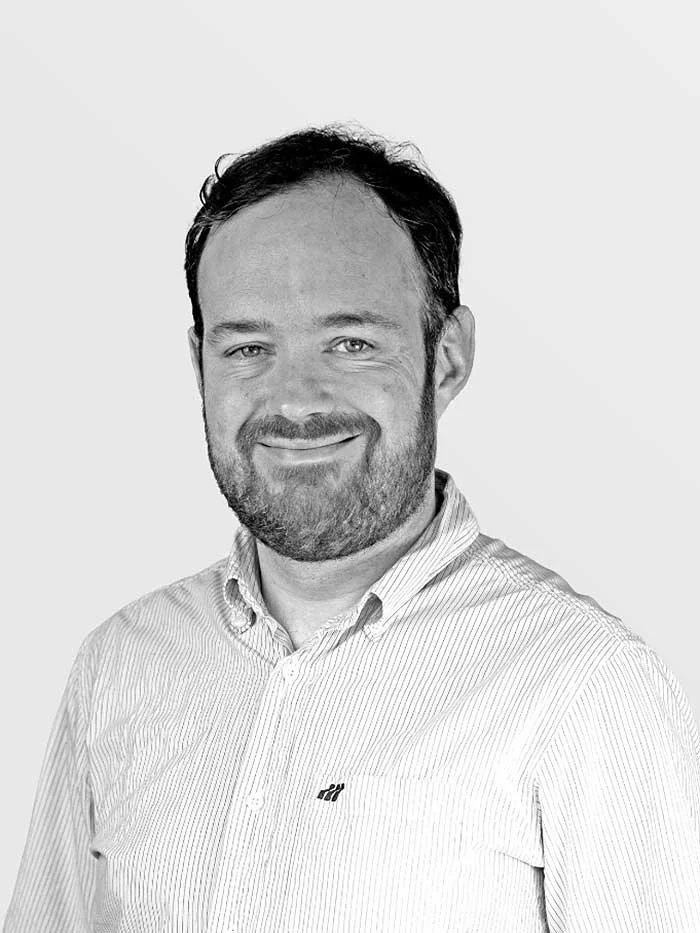 Black and white headshot of a smiling man with a beard and short dark hair, wearing a striped button-up shirt, against a plain background.