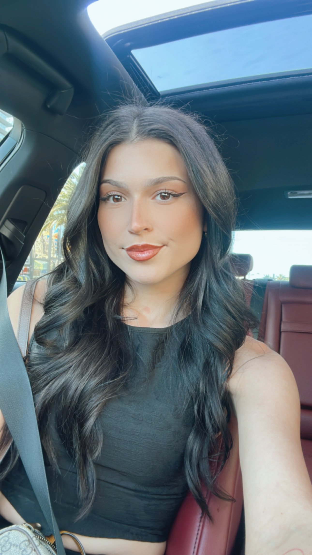 A young woman with long, wavy dark hair, makeup, and a black sleeveless top taking a selfie inside a car with red leather seats and a sunroof.
