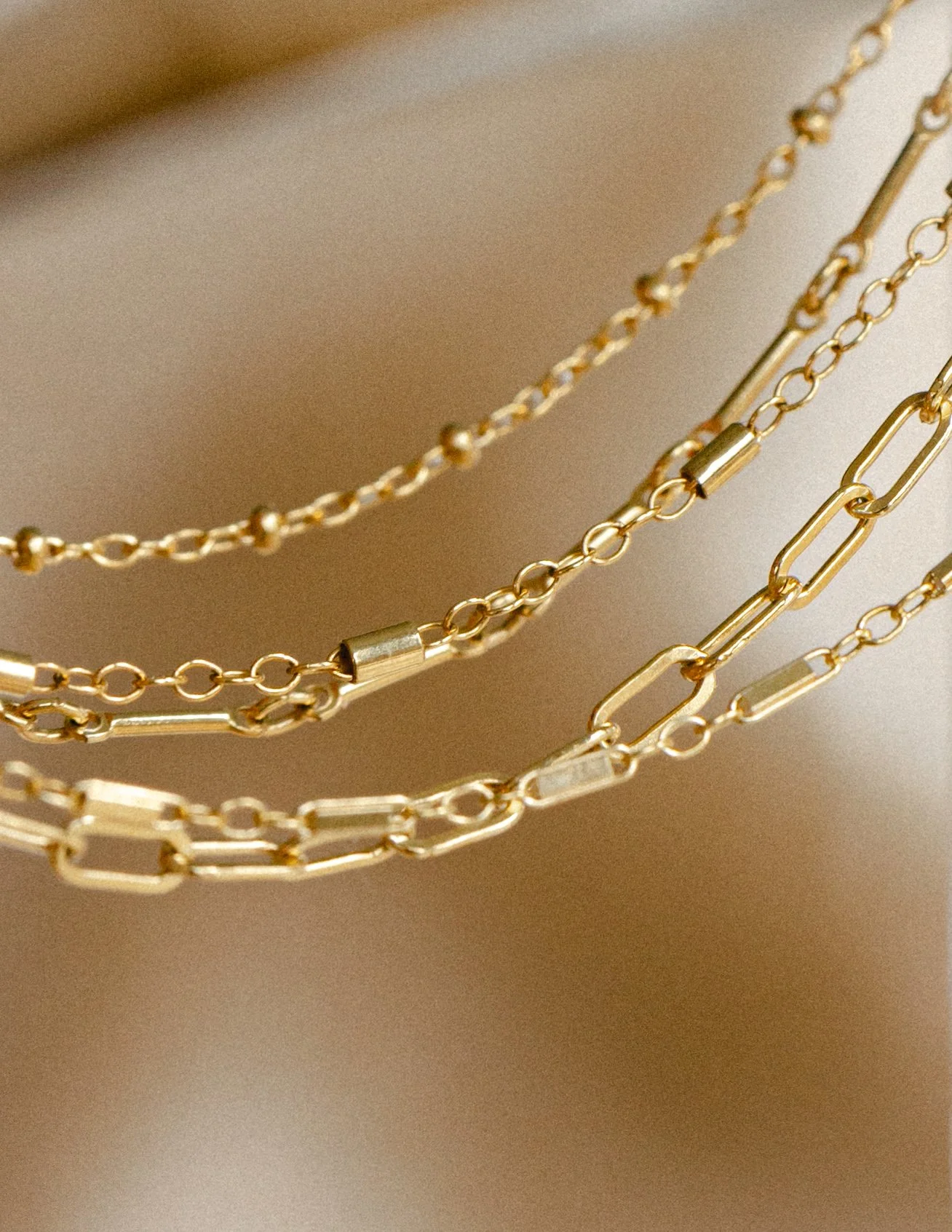 Close-up of four gold layered necklaces with different chain styles, hanging against a soft beige background.