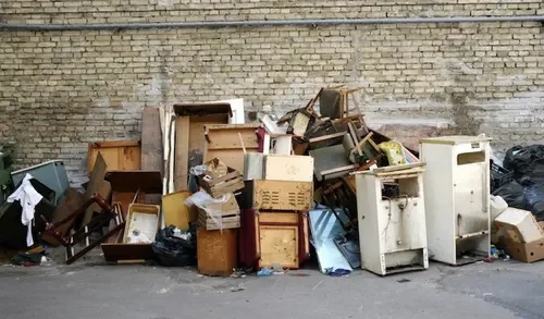 Pile of discarded furniture and appliances, including chairs, dressers, and a mini refrigerator, against a brick wall.