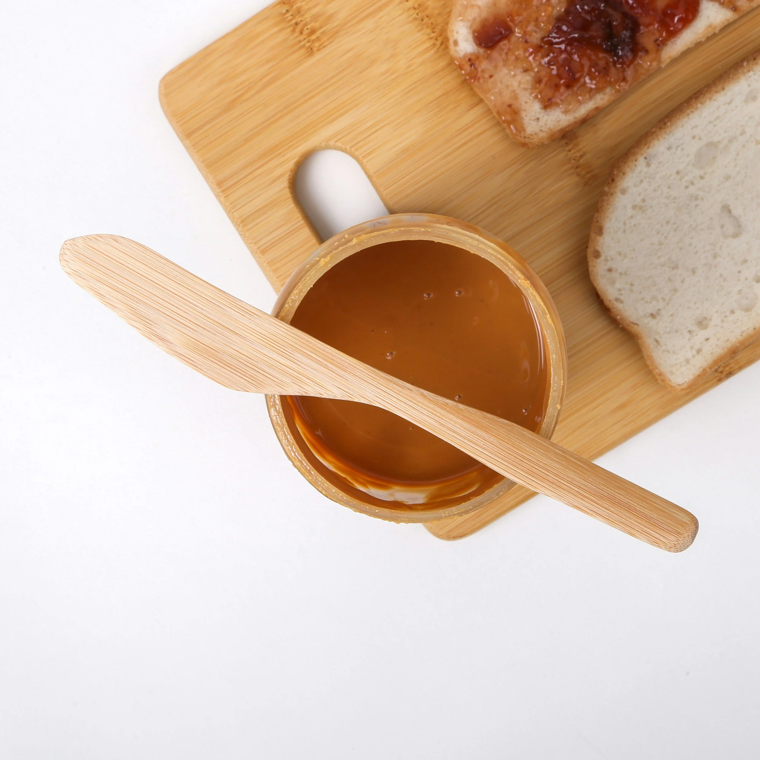 Peanut butter jar with a wooden spreader, sliced bread, and a piece of toast with jam on a wooden cutting board.