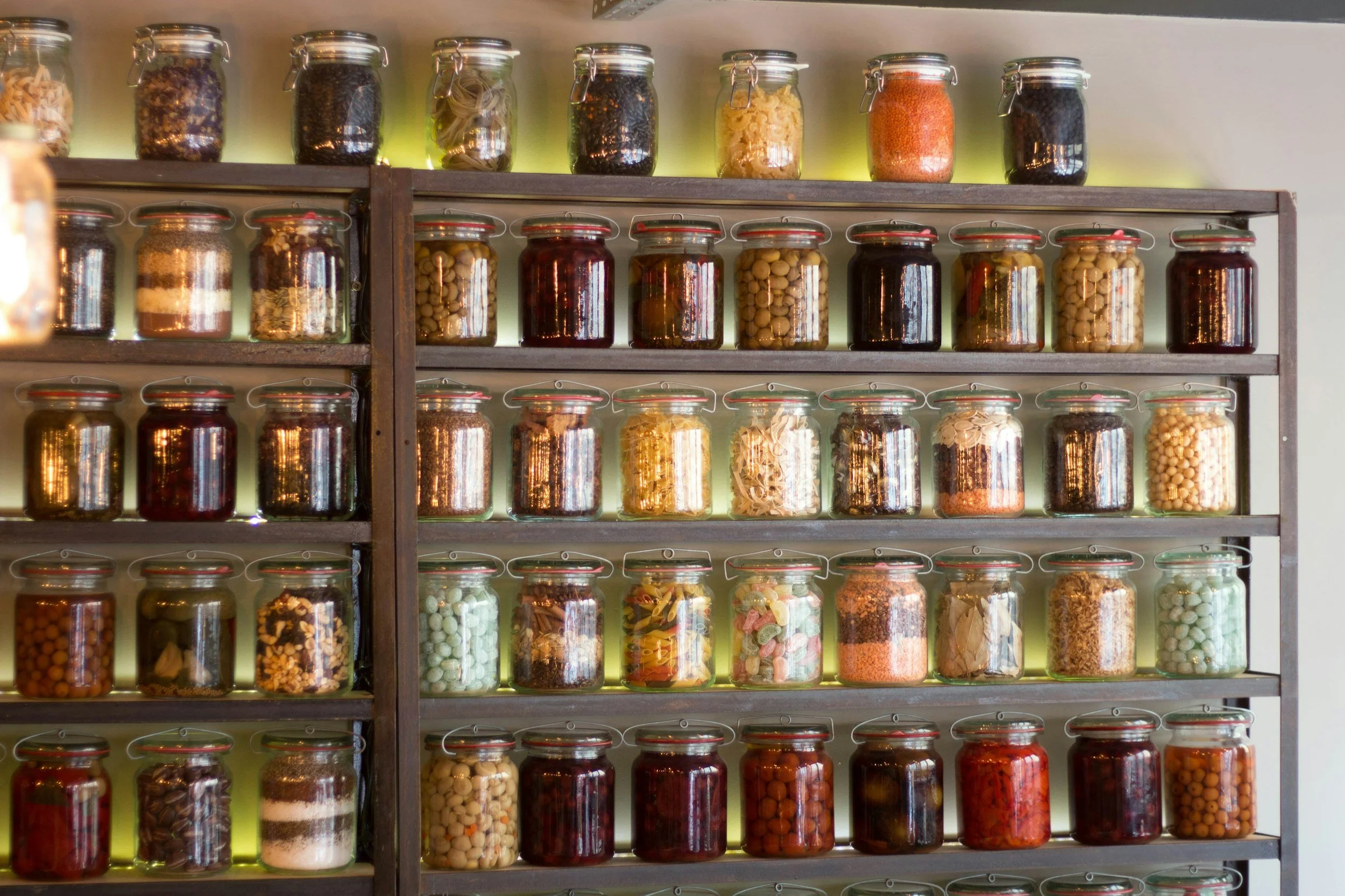 A large wooden shelf filled with glass jars containing various spices, herbs, and dried goods, with some jars illuminated from behind.