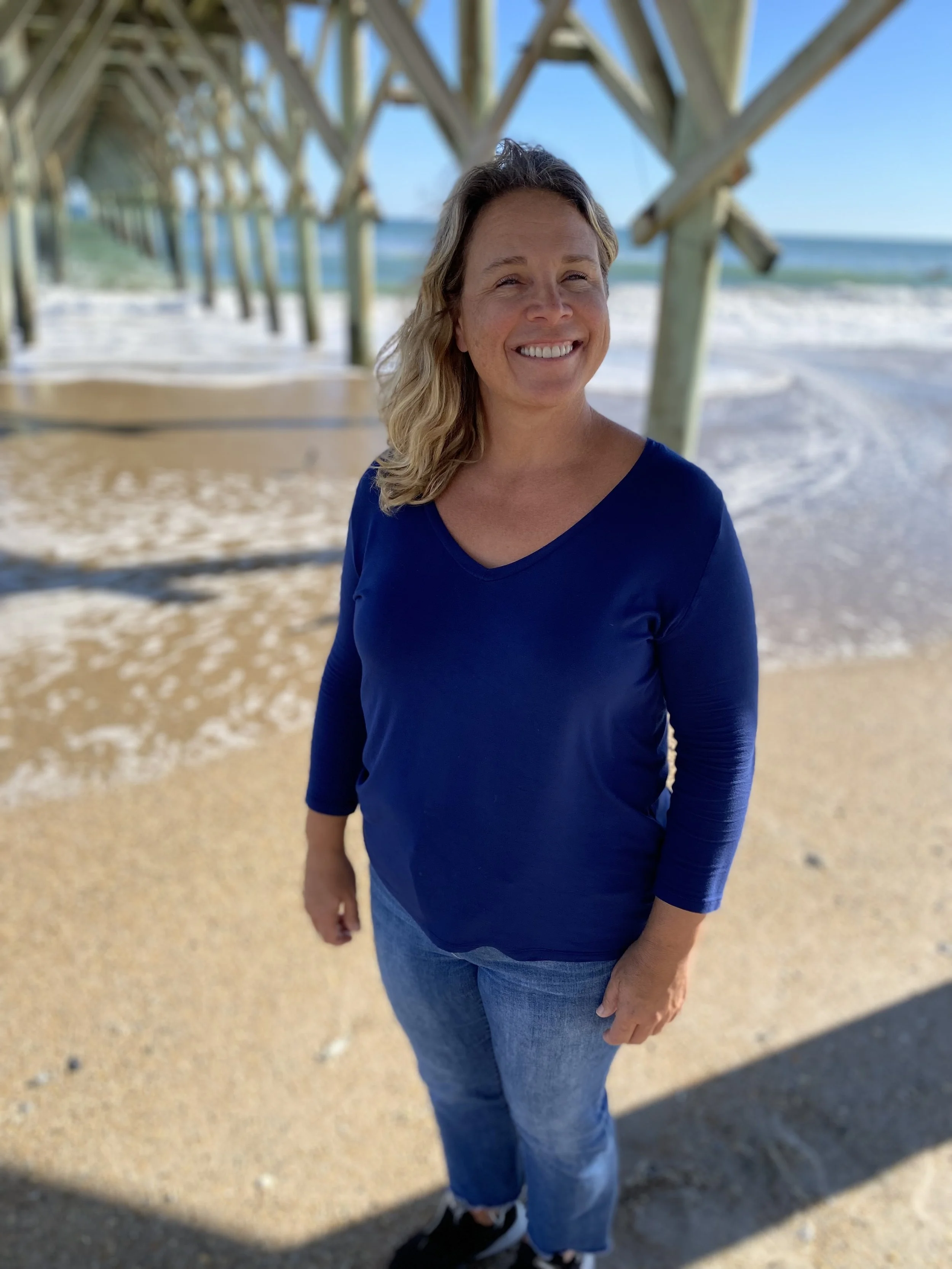 Woman standing on a sandy beach under a wooden pier, smiling, wearing a navy blue long-sleeve top and jeans.
