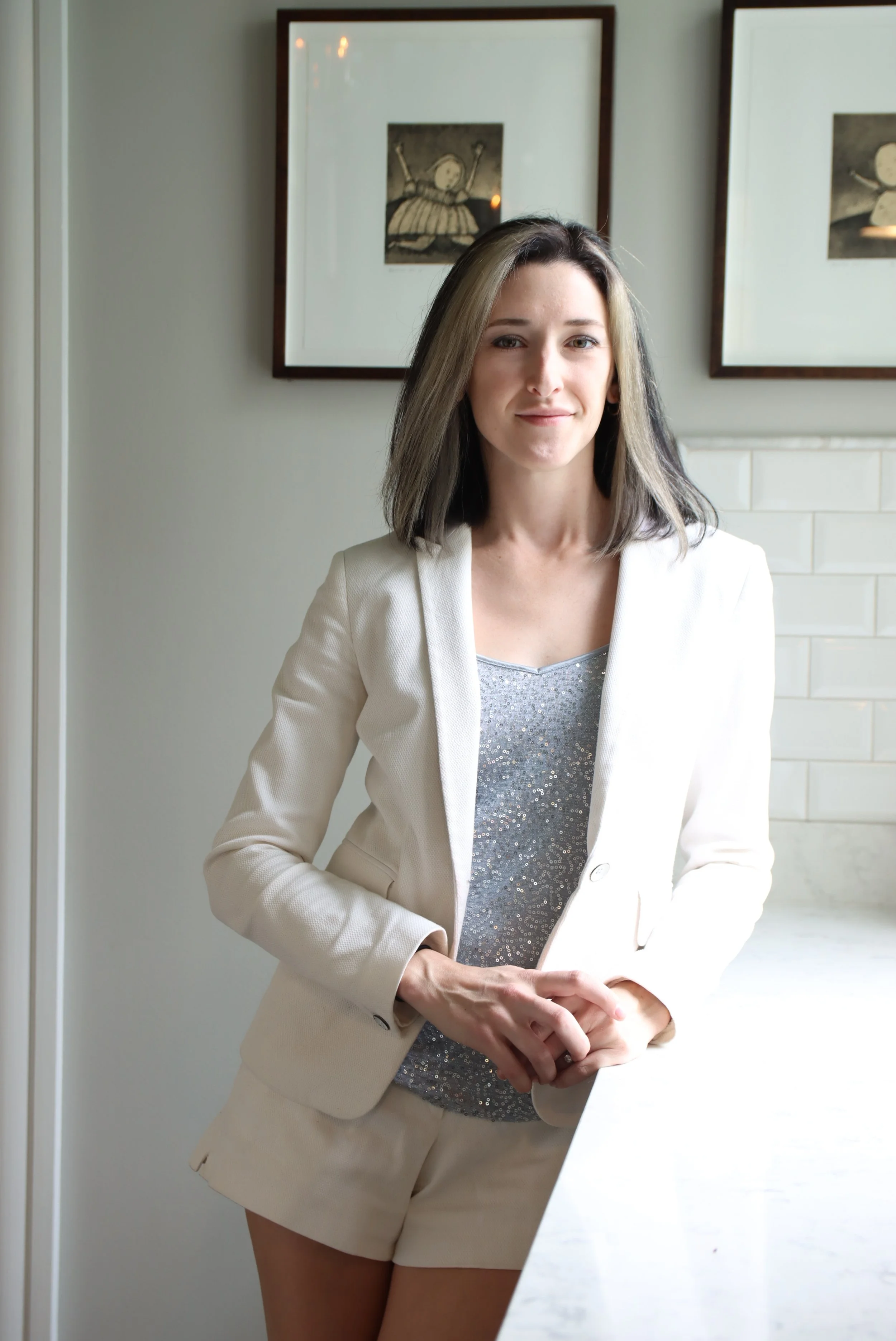 A woman with shoulder-length dark hair and light highlights, wearing a white blazer over a sparkly silver top and matching shorts, standing indoors in front of framed artwork and a white tiled wall.