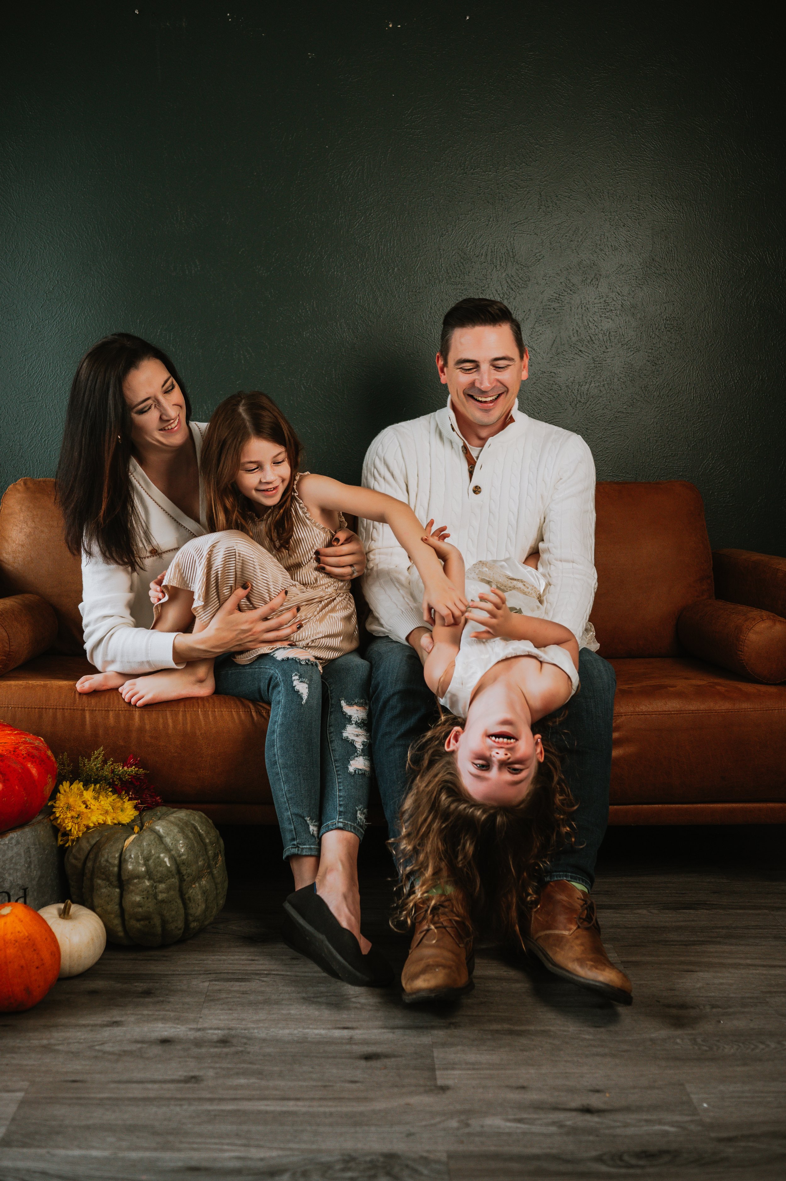 A family of five enjoying time together on a brown couch with pumpkins and flowers on the floor nearby, with a dark green wall in the background.