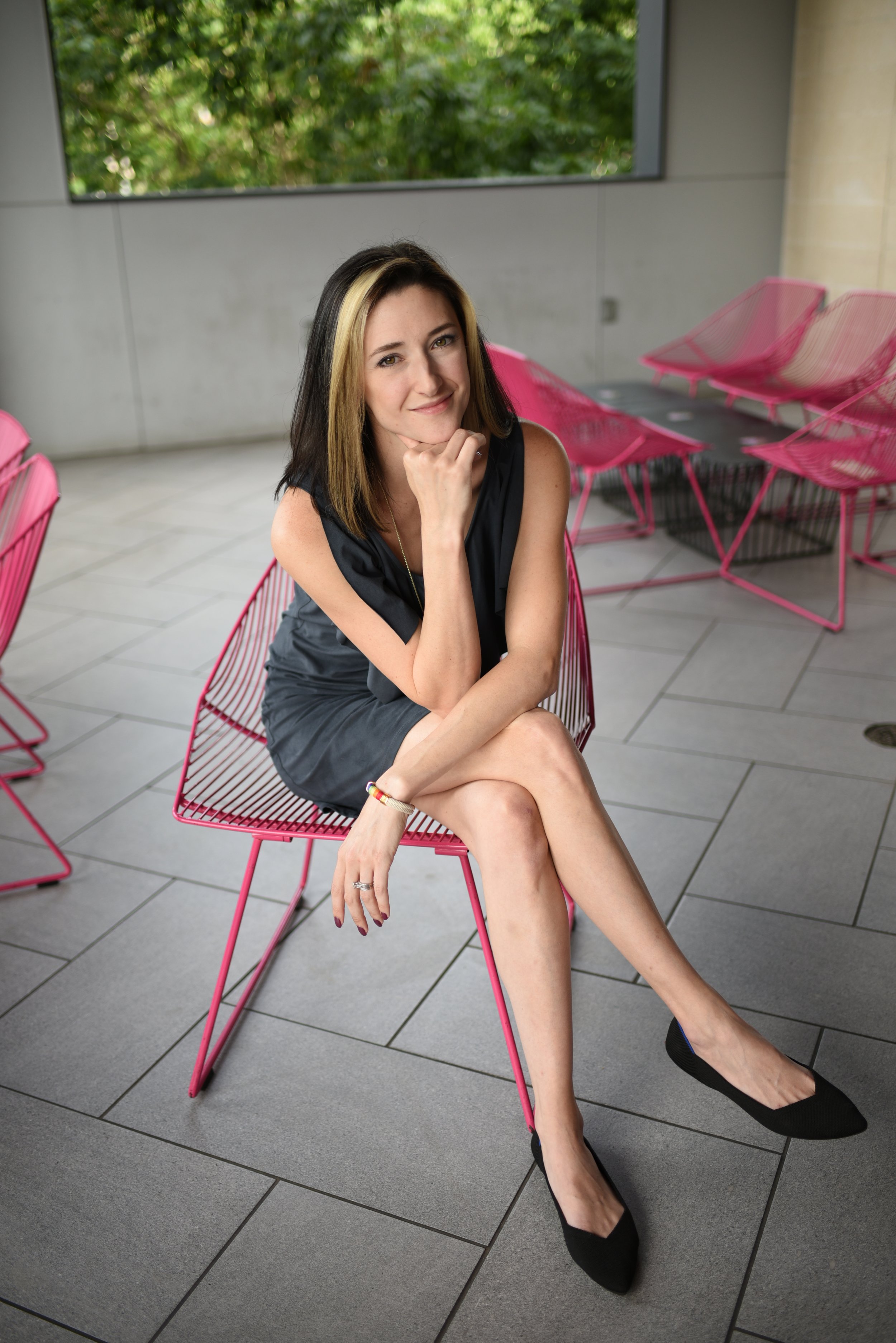 A woman sitting on a pink wire chair with her legs crossed, wearing a black dress and black flats, smiling and resting her chin on her hand in a modern indoor space with large windows and pink chairs in the background.