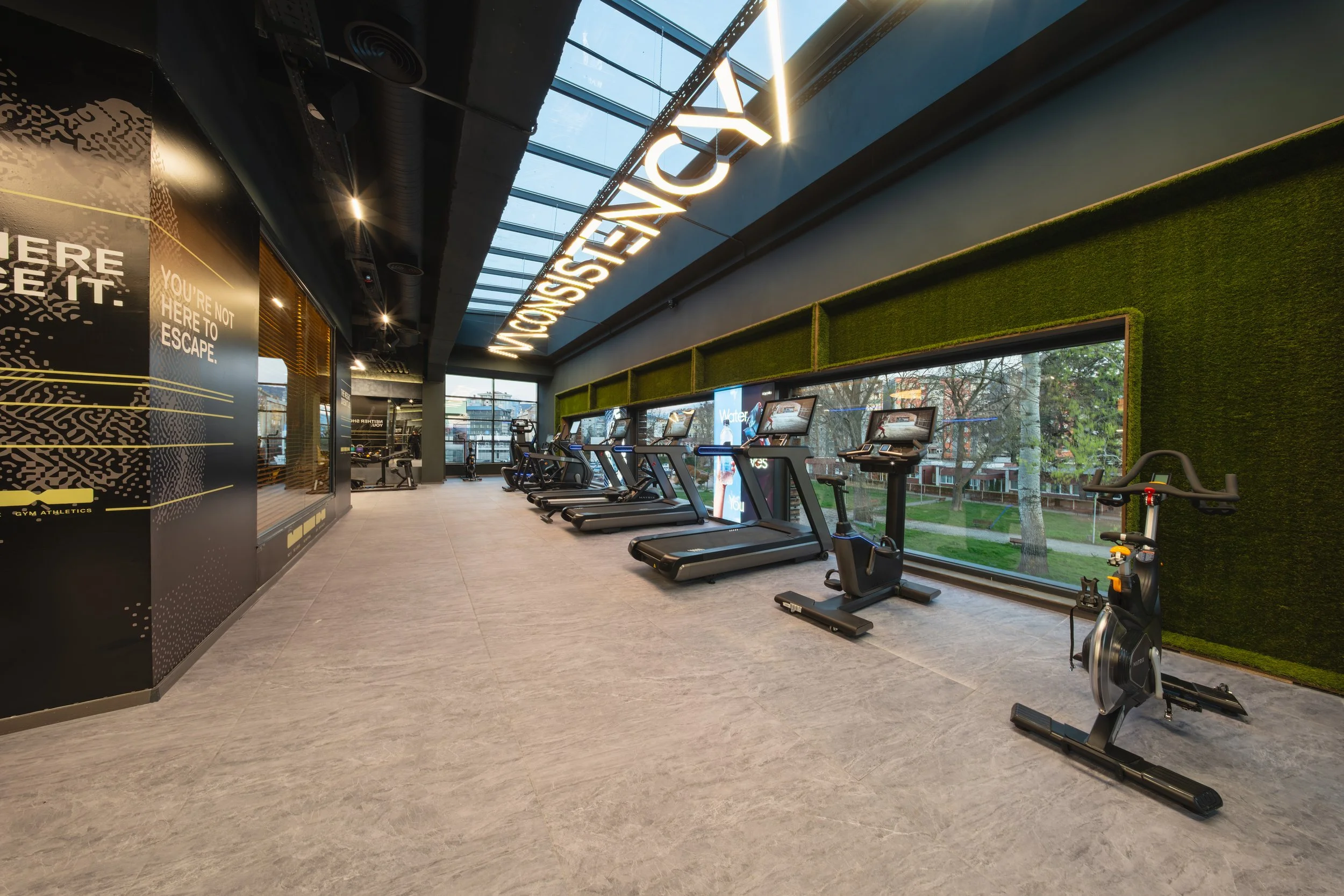 Interior of a modern gym with cardio equipment such as treadmills and exercise bikes, large windows showing trees outside, and decorative green wall panels.