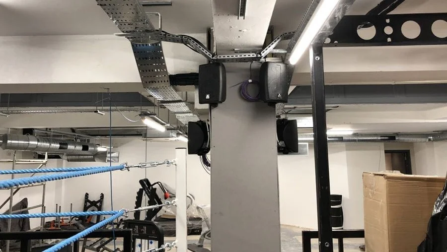 Photo of an indoor gym with black speakers, a punching bag, and blue boxing ropes.