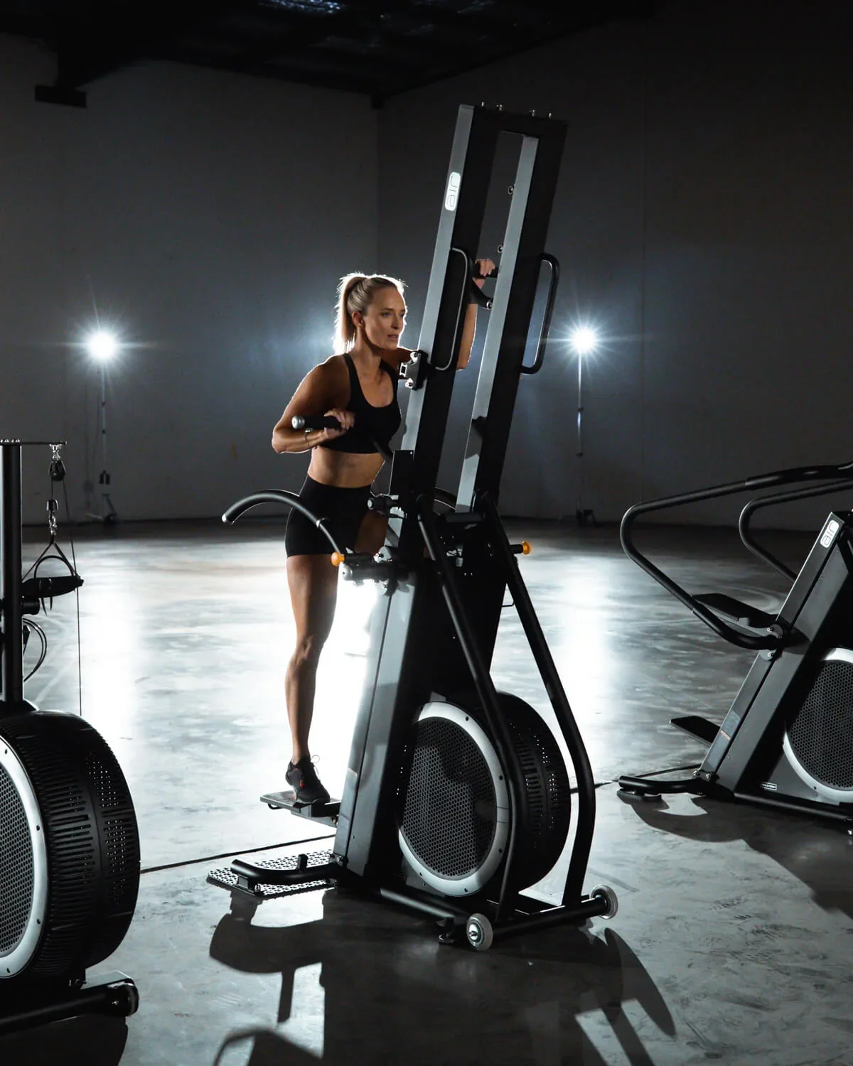 A woman in black workout clothes doing a step exercise on a steppers machine in a gym with dark walls and bright backlighting.