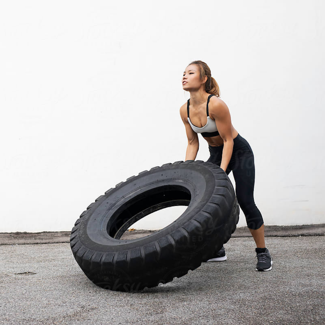 Woman in athletic wear flipping a large black tire outdoors.