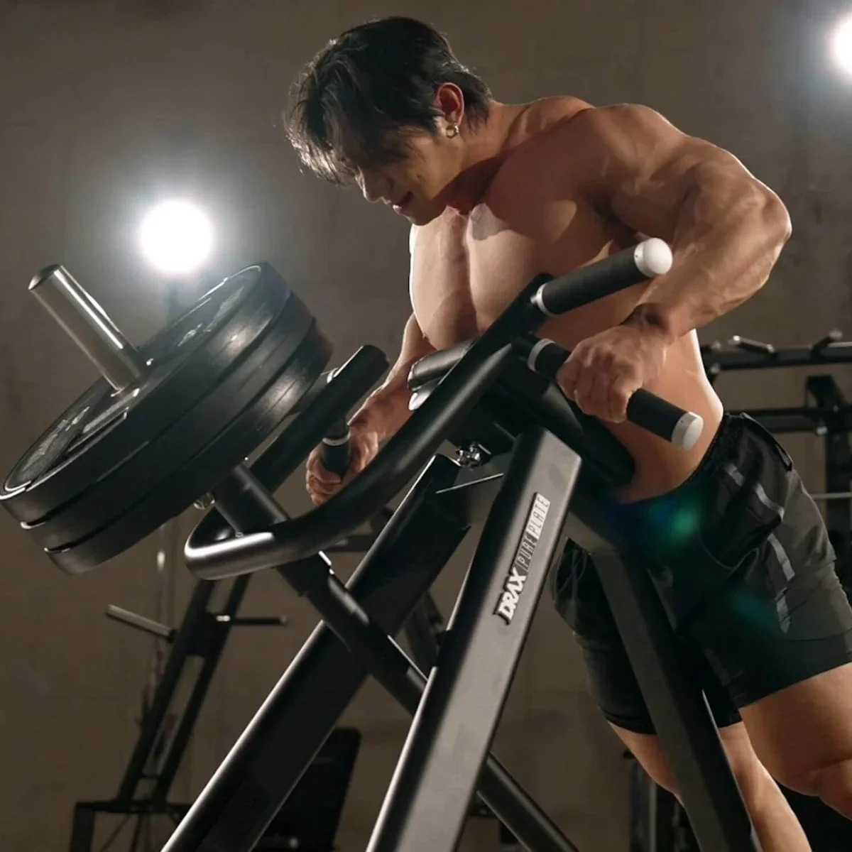 A muscular man performing a workout on a piece of gym equipment with weights, bent forward and gripping the handles.