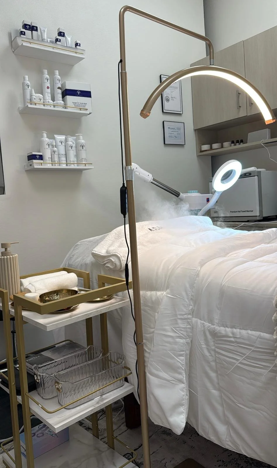 A spa or treatment room with a bed covered in white linens, facial tools on a gold cart, shelves with skincare products, a magnifying lamp, and a steam or vapor device emitting mist near the bed.