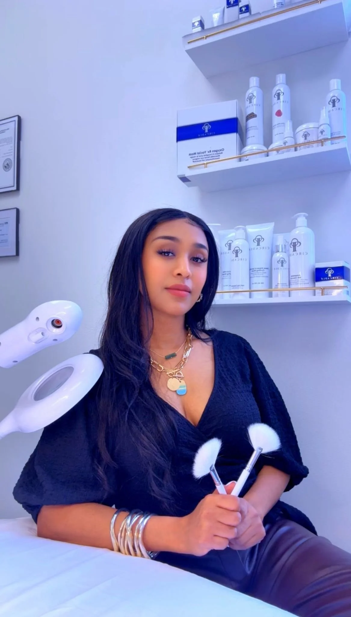 A woman sitting on a bed in a spa or beauty clinic, holding facial brushes in her hands, with skincare products on shelves behind her.