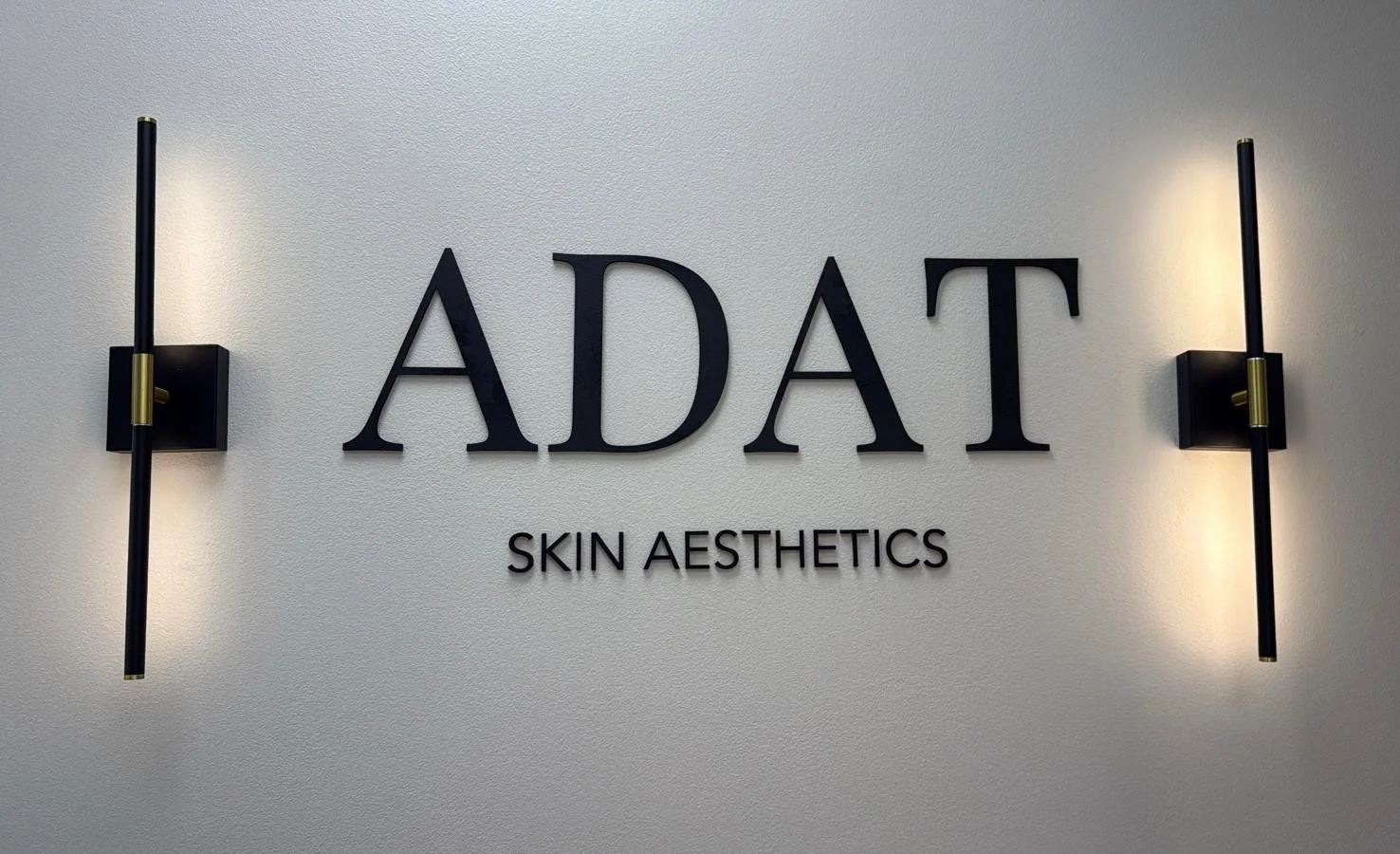 Sign reading 'A DAT Skin Aesthetics' with wall-mounted lights on a textured wall