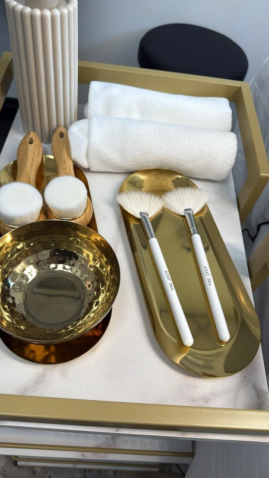 Luxury skincare tools and accessories on a gold tray and table, including white towels, makeup brushes labeled 'Skincare', a gold bowl, and a gold tray with cotton pads and skincare spatulas.