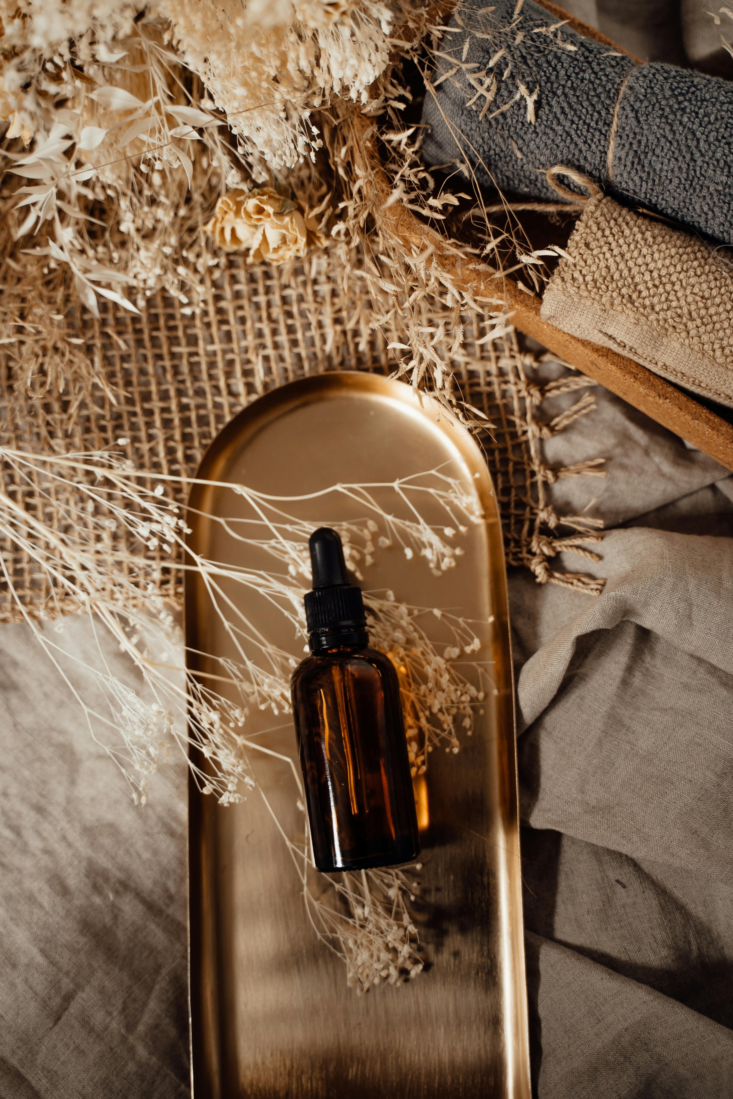 An amber glass dropper bottle placed on a metallic tray with dried beige flowers around it, surrounded by folded fabrics in neutral tones.