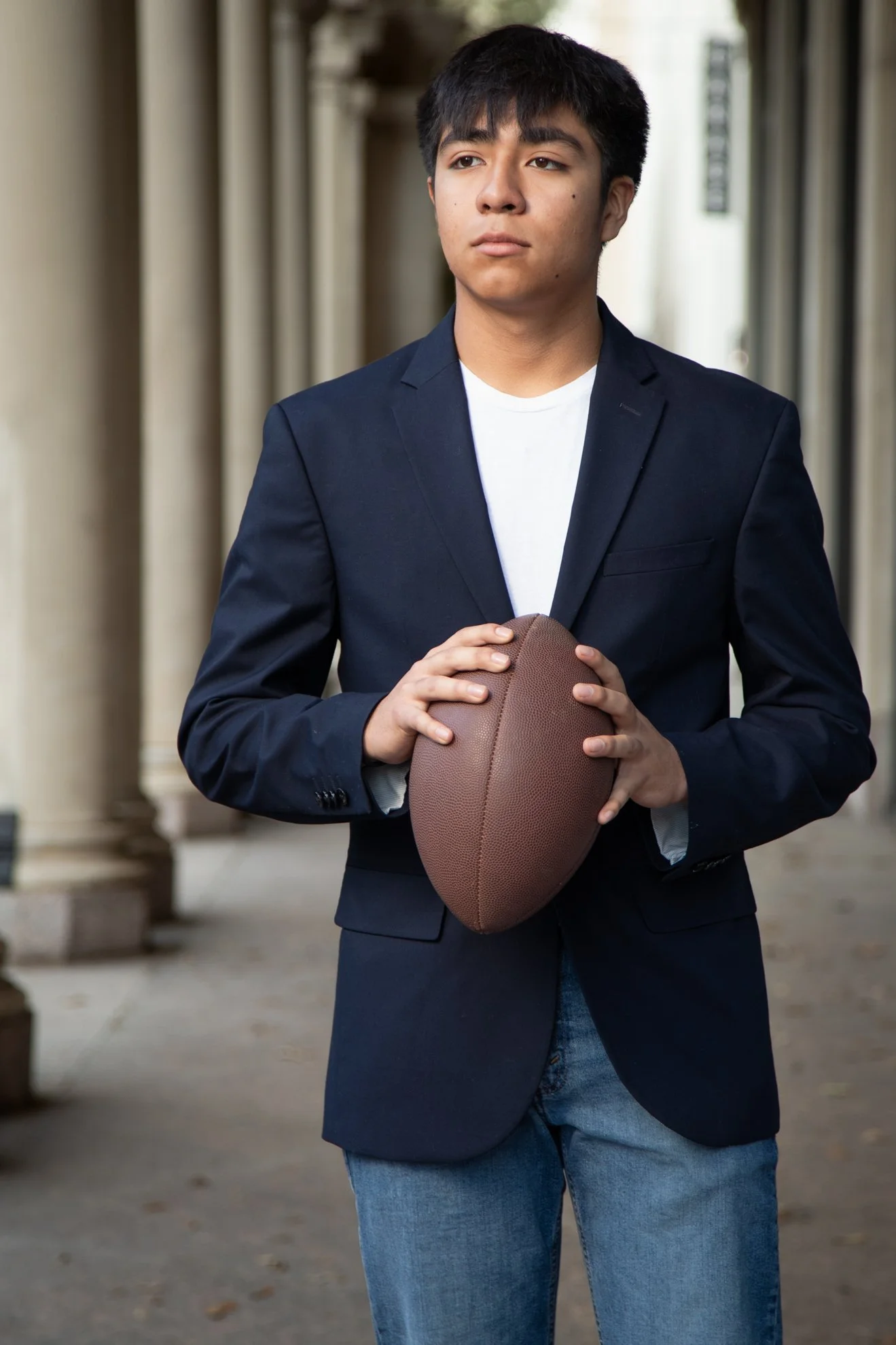 A Houston high school senior young man in a navy blazer and jeans holding a football, standing outdoors near columns.